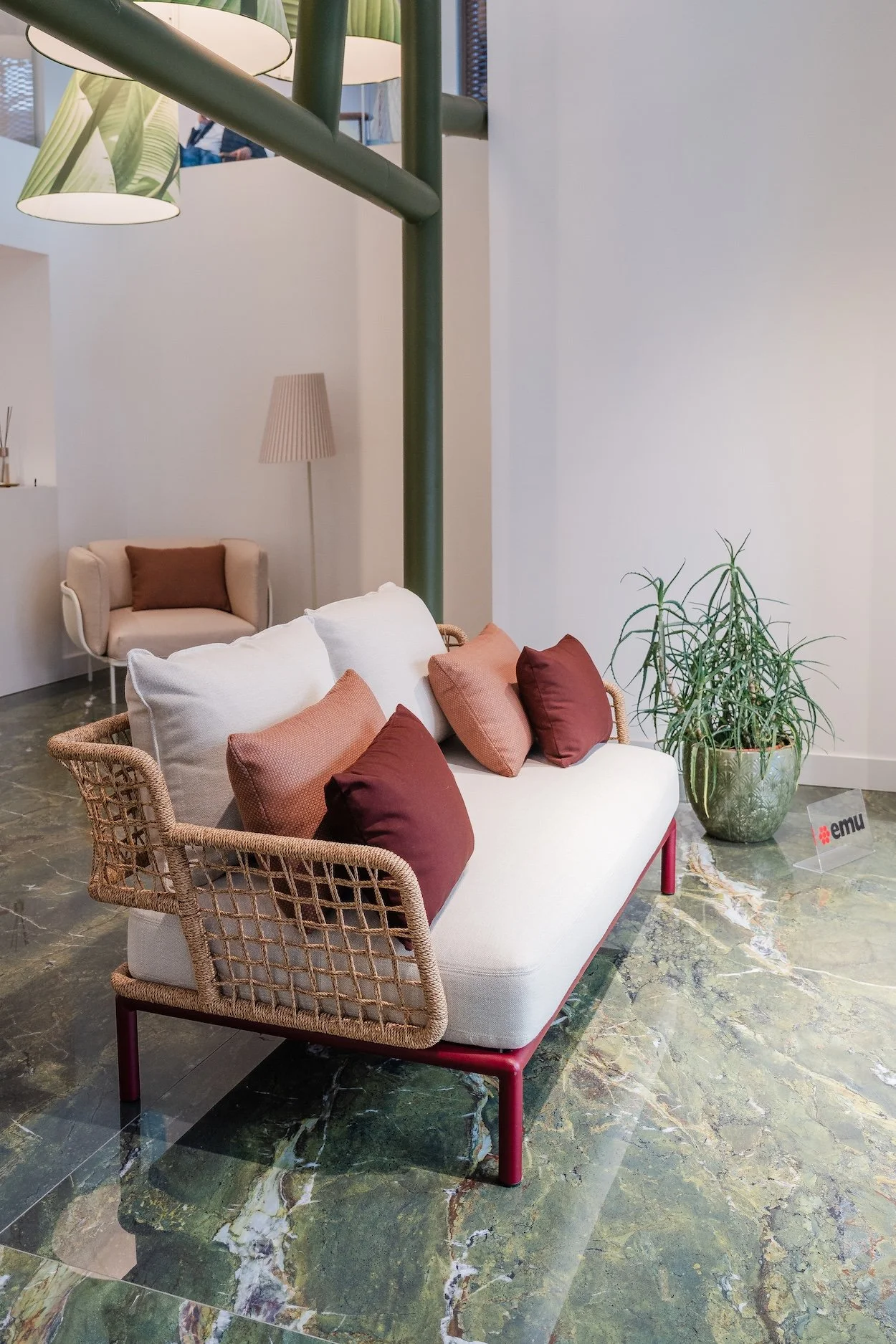 Modern lounge area with a white cushioned sofa accented with red and brown pillows, a beige armchair with a brown pillow, a green potted plant, and a tall floor lamp, on a marble floor.