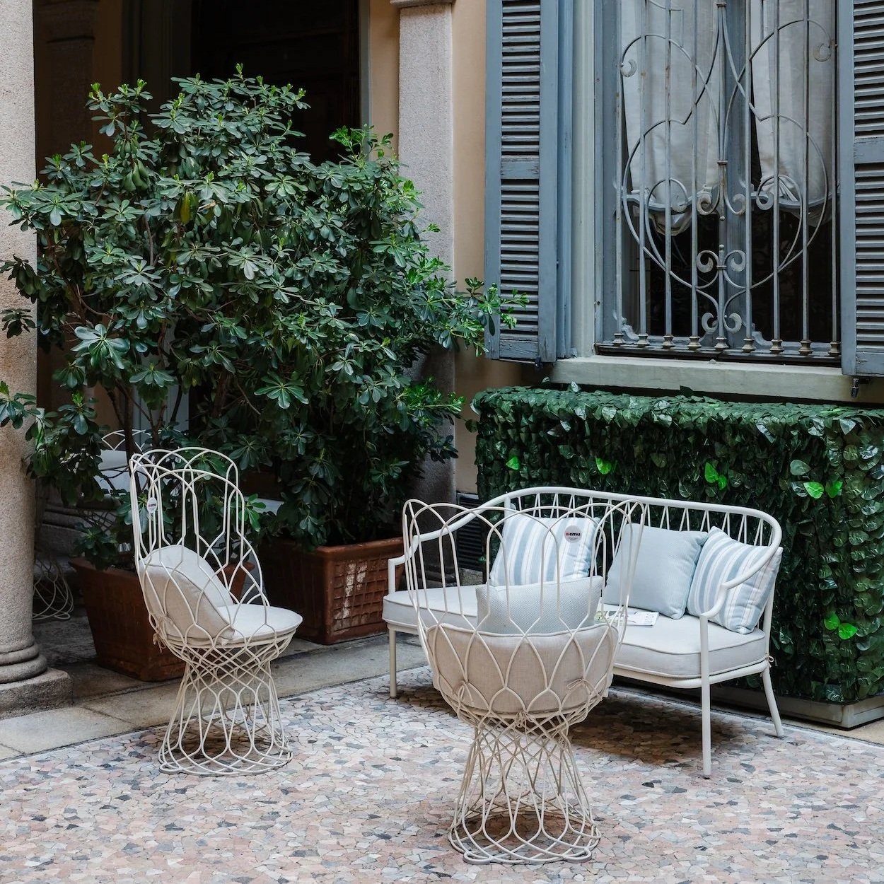Outdoor patio with white wire chairs, a white cushioned settee, pillows, potted plants, and a hedge in the background.