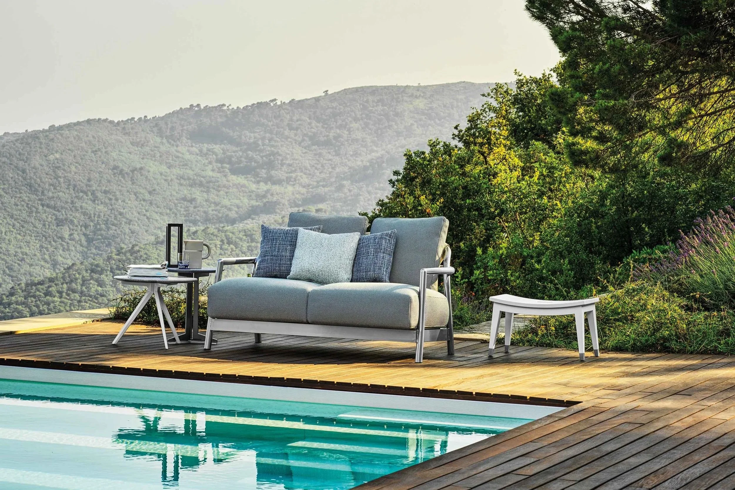 Outdoor patio with a lounge chair, side table, and a small white stool beside a swimming pool, with lush green trees and mountain landscape in the background.