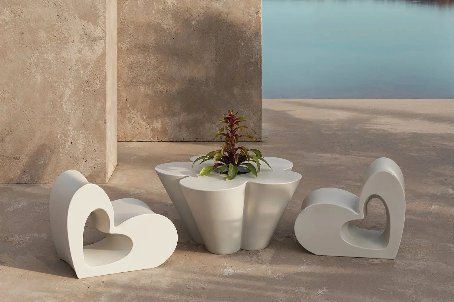 Two white heart-shaped chairs and a white flower pot with a plant, set on a concrete surface near water with a large stone structure in the background.