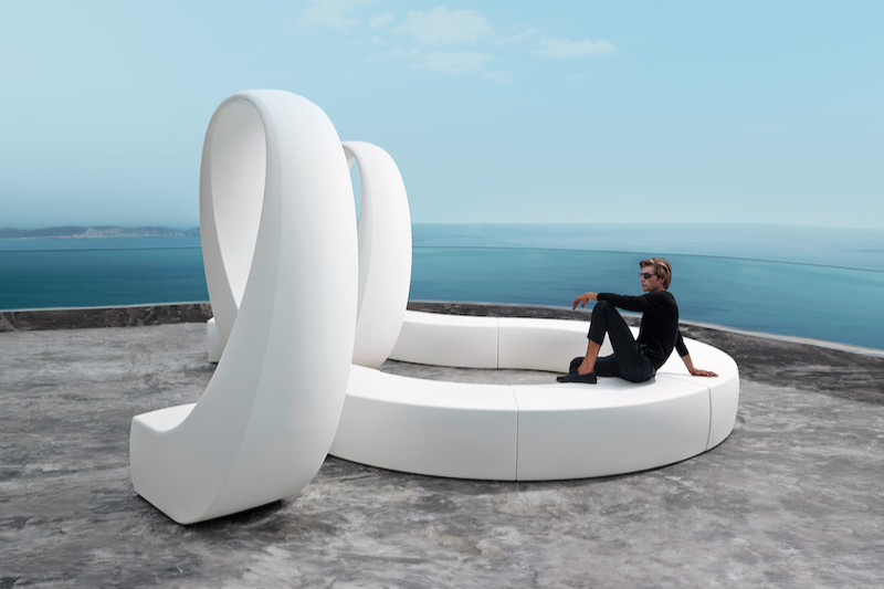 A woman sitting on a modern white sculpture shaped like a hand overlooking the ocean.