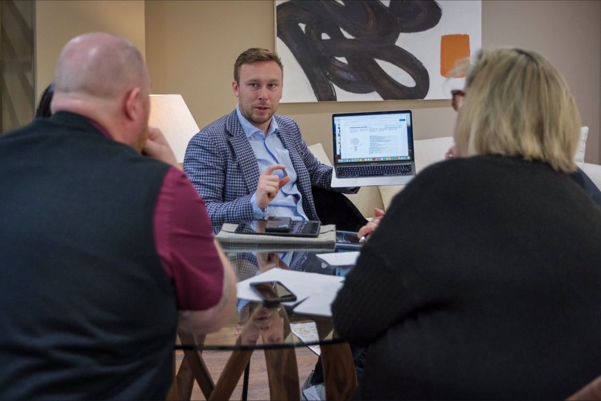 A group of four people having a business meeting around a glass table, with a man showing something on a laptop.