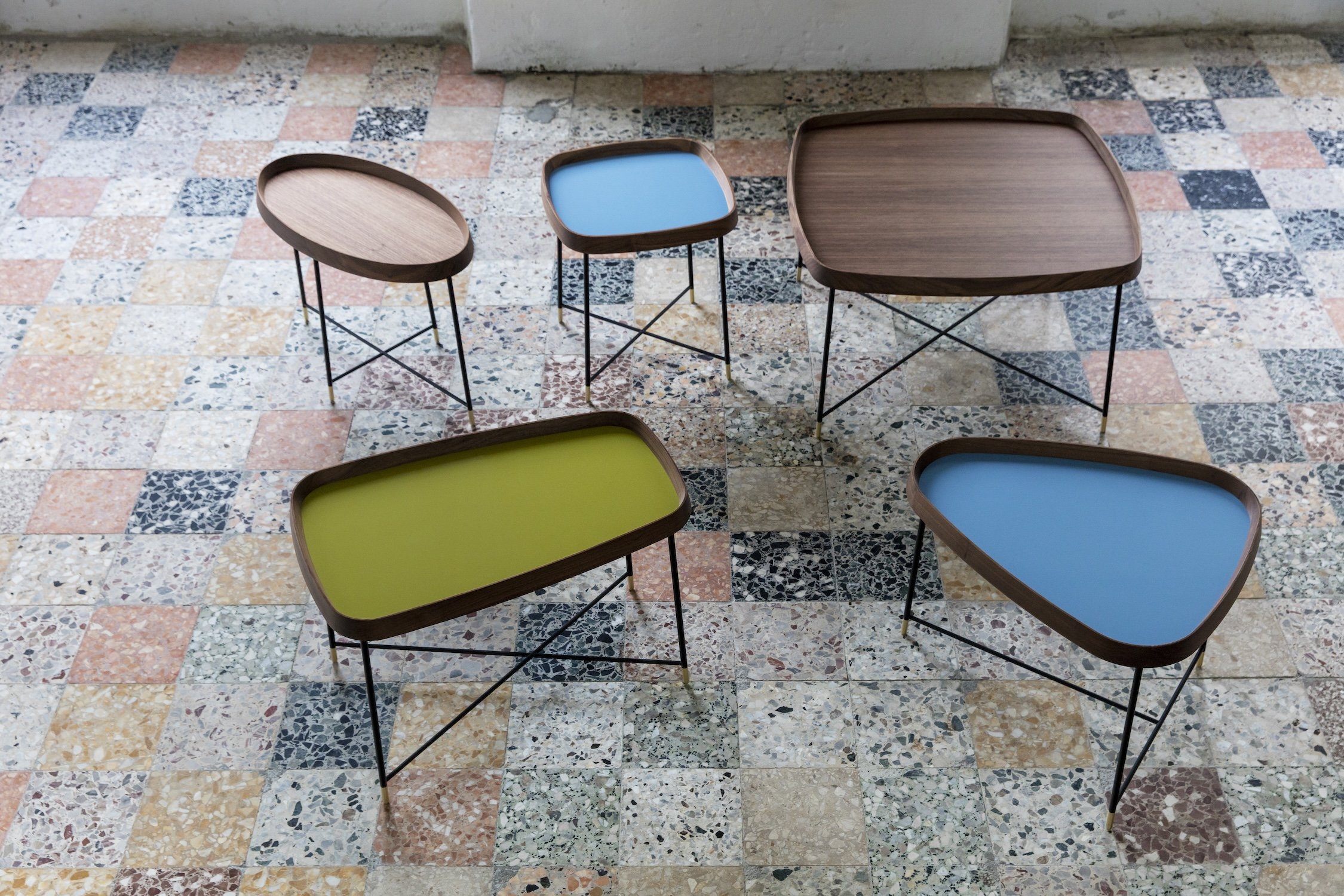 Six modern wooden tables with metal legs are placed on a colorful tiled floor. The tables have different shapes and colors: one with a blue top, one with a green top, one with a square brown top, one with an oval brown top, and two with round brown t