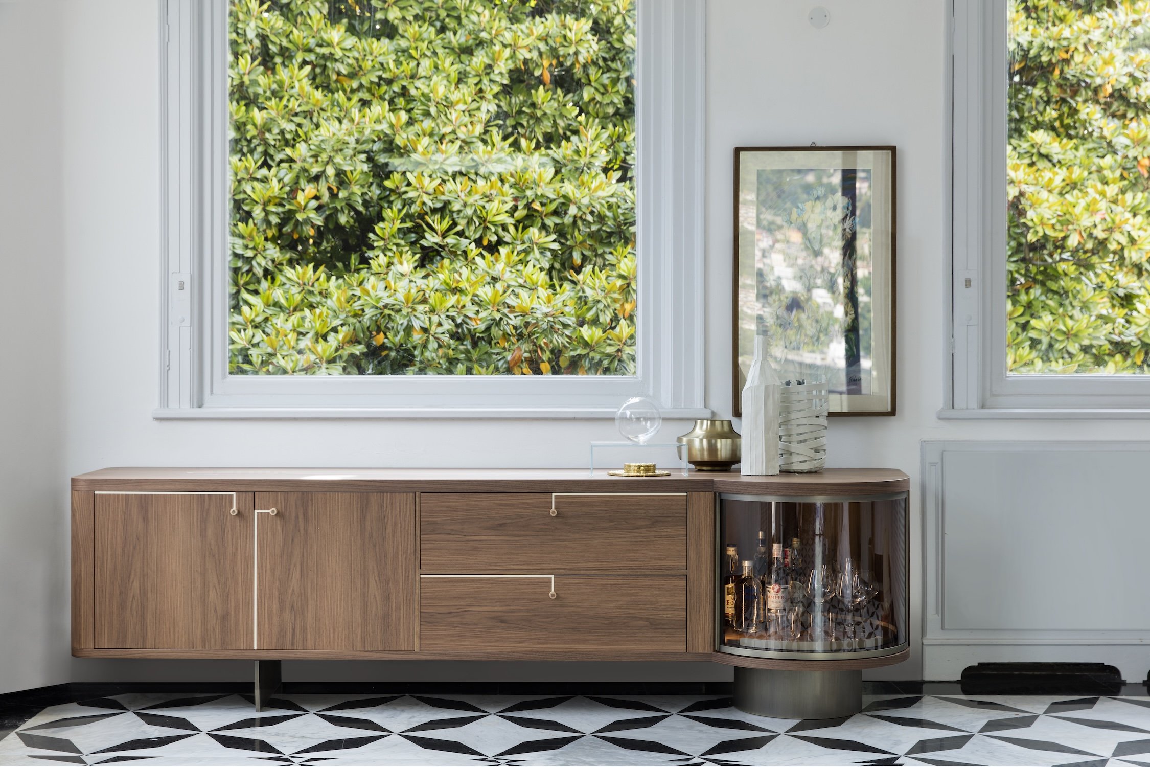 A modern wooden sideboard with decorative items on top, positioned in front of large windows overlooking a lush green tree. The sideboard has a curved glass cabinet section with bottles inside and is mounted on a black-and-white geometric patterned f