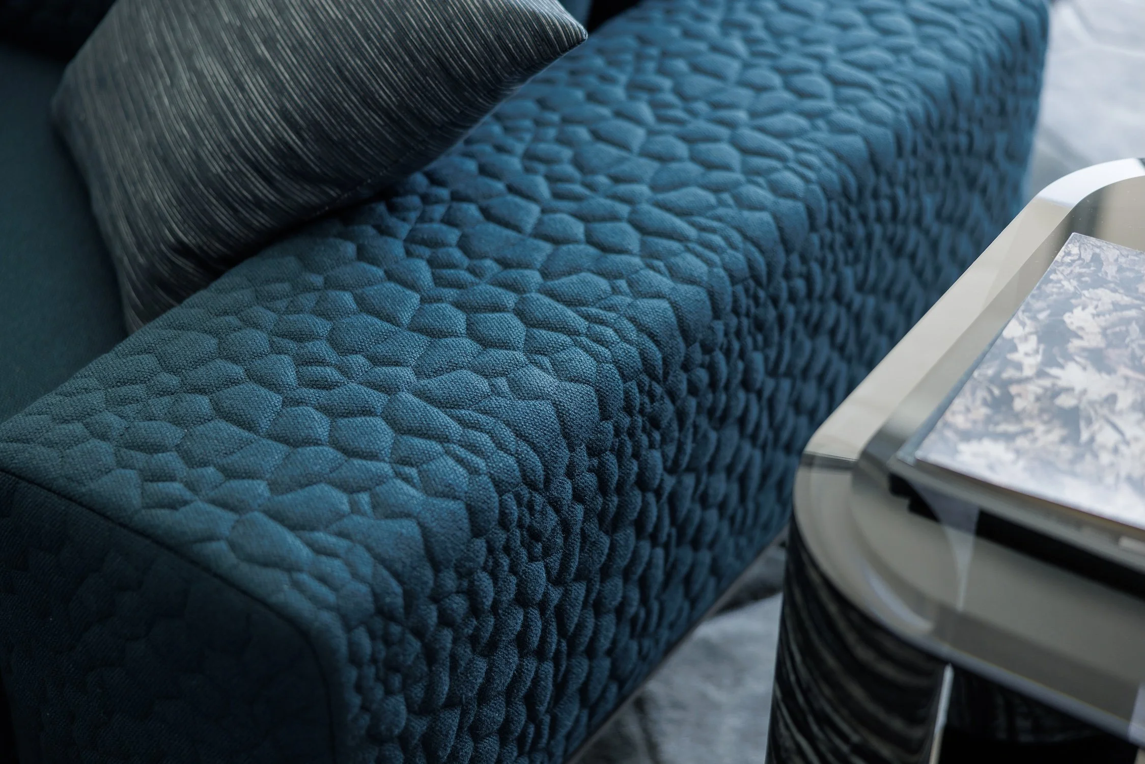 Close-up of a textured dark blue upholstered couch with a matching throw pillow, and a glass-topped side table with a reflective surface beside it.