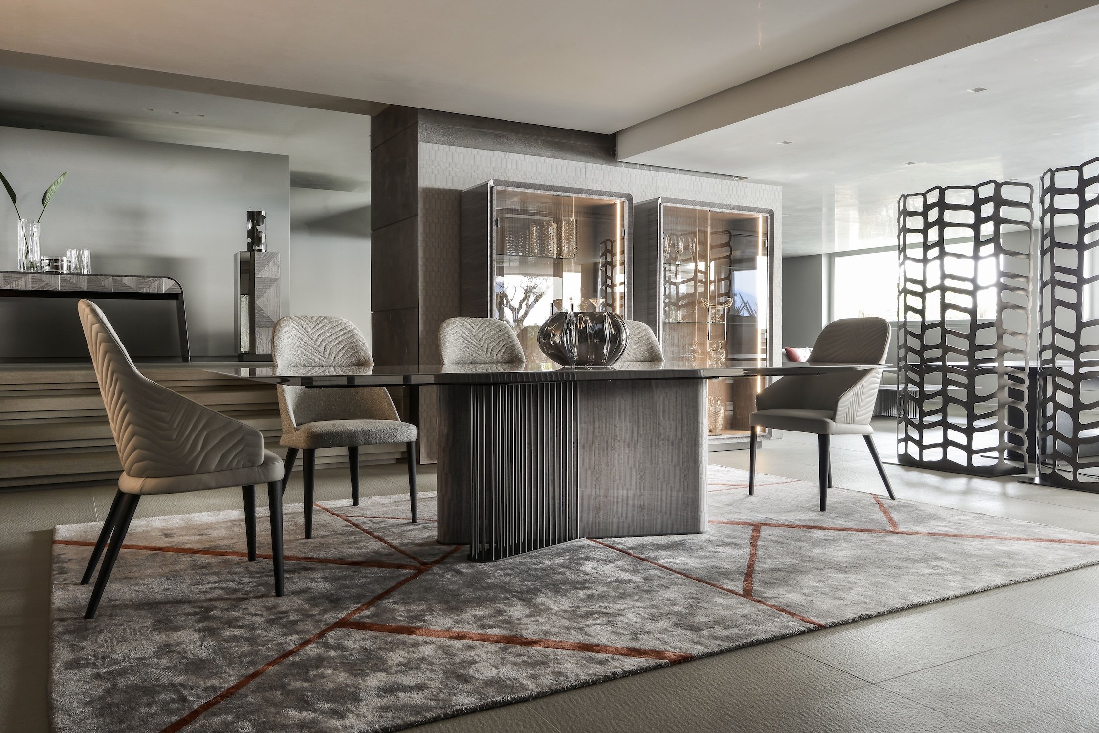 Modern dining room with a rectangular glass table, four upholstered chairs, a patterned rug, and decorative display cabinets in the background.