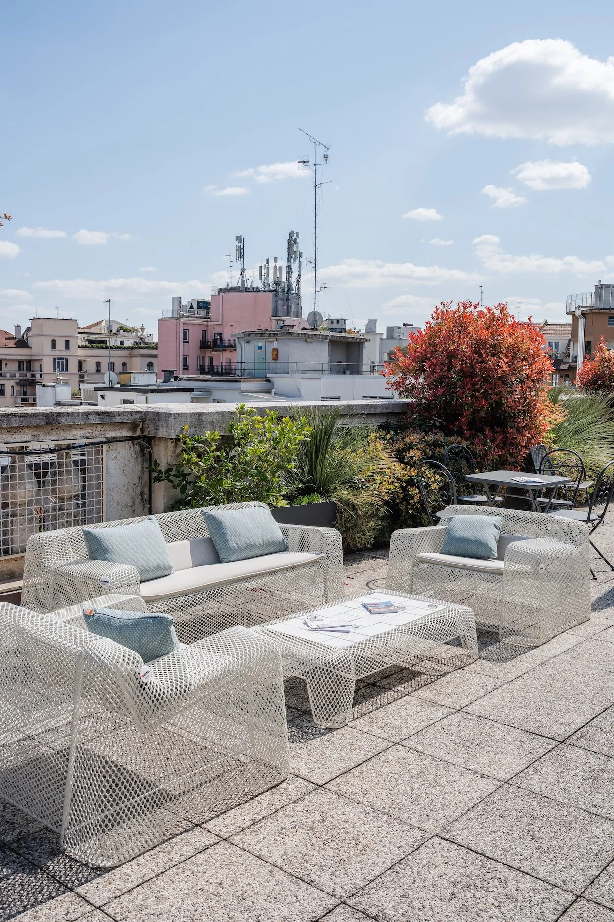 Urban rooftop terrace with modern white furniture including a sofa, armchairs, and a coffee table, surrounded by colorful plants and view of city buildings and antennas under a partly cloudy sky.