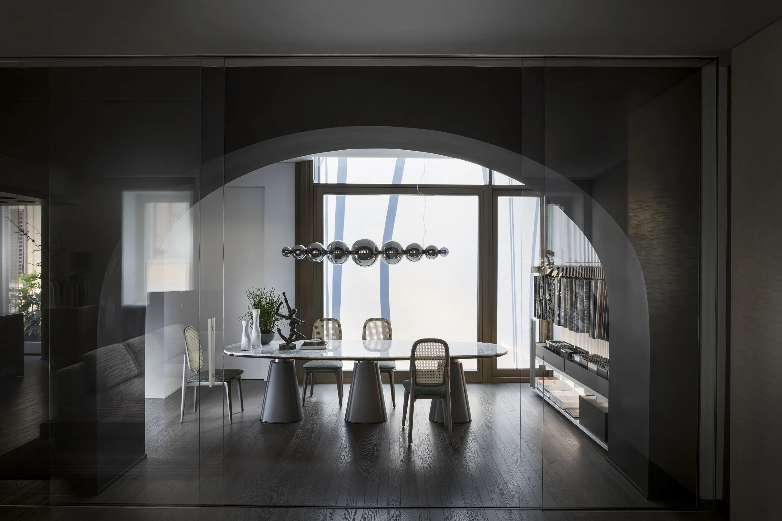 Modern dining room with a marble table, six chairs, a large window, a black chandelier, plants, sculptures, and a bookshelf, with a dark wood floor.