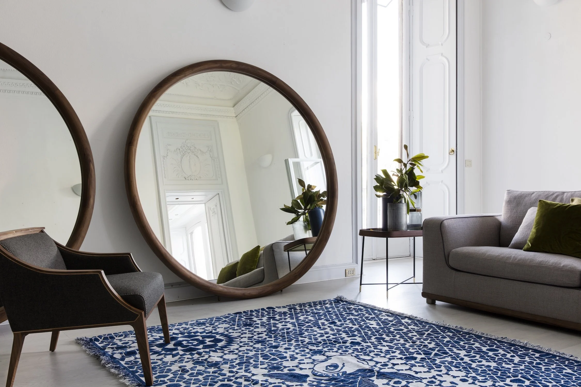 Interior of a modern living room with a large round mirror reflecting an ornate ceiling, a grey armchair, a grey sofa with green pillows, a small table with potted plants, and a patterned blue and white rug on a light-colored floor.
