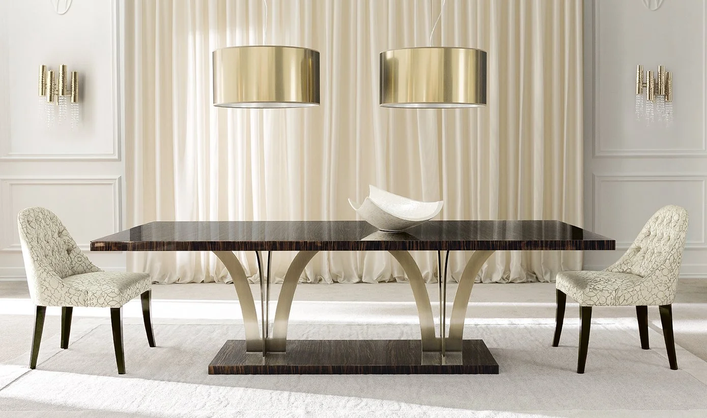 Modern dining room with a dark wood table, two cream upholstered chairs, hanging gold pendant lights, white curtains, and a white decorative bowl on the table.