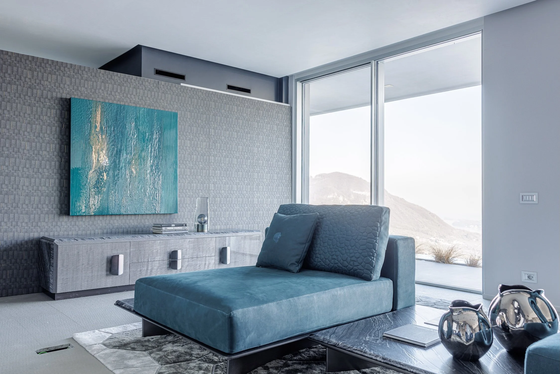 Modern living room with blue velvet chaise lounge, textured gray wall, large abstract ocean painting, and floor-to-ceiling windows overlooking a mountain landscape.
