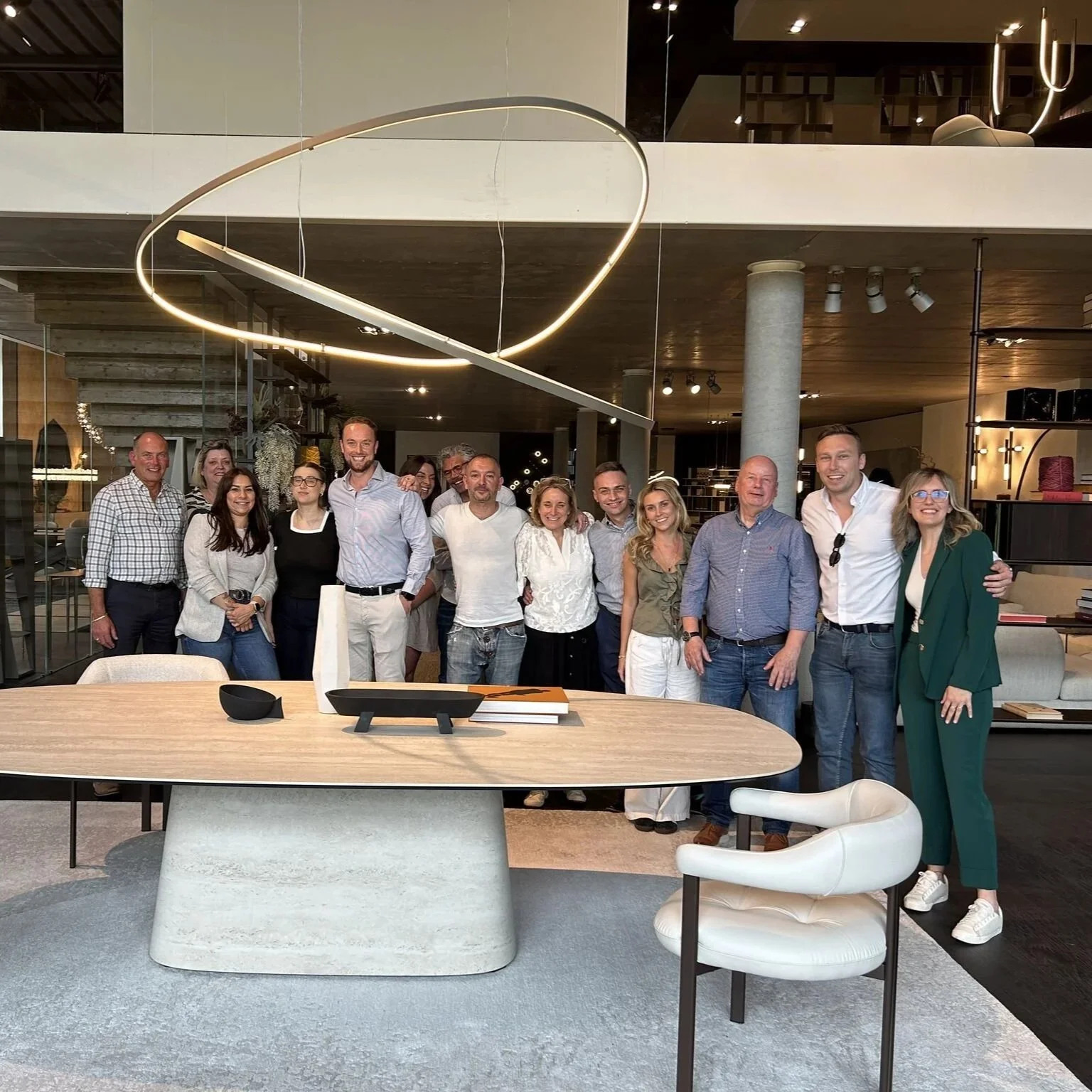A group of fifteen people standing inside a modern, stylish room with contemporary lighting, posing for a photo around a large wooden table.