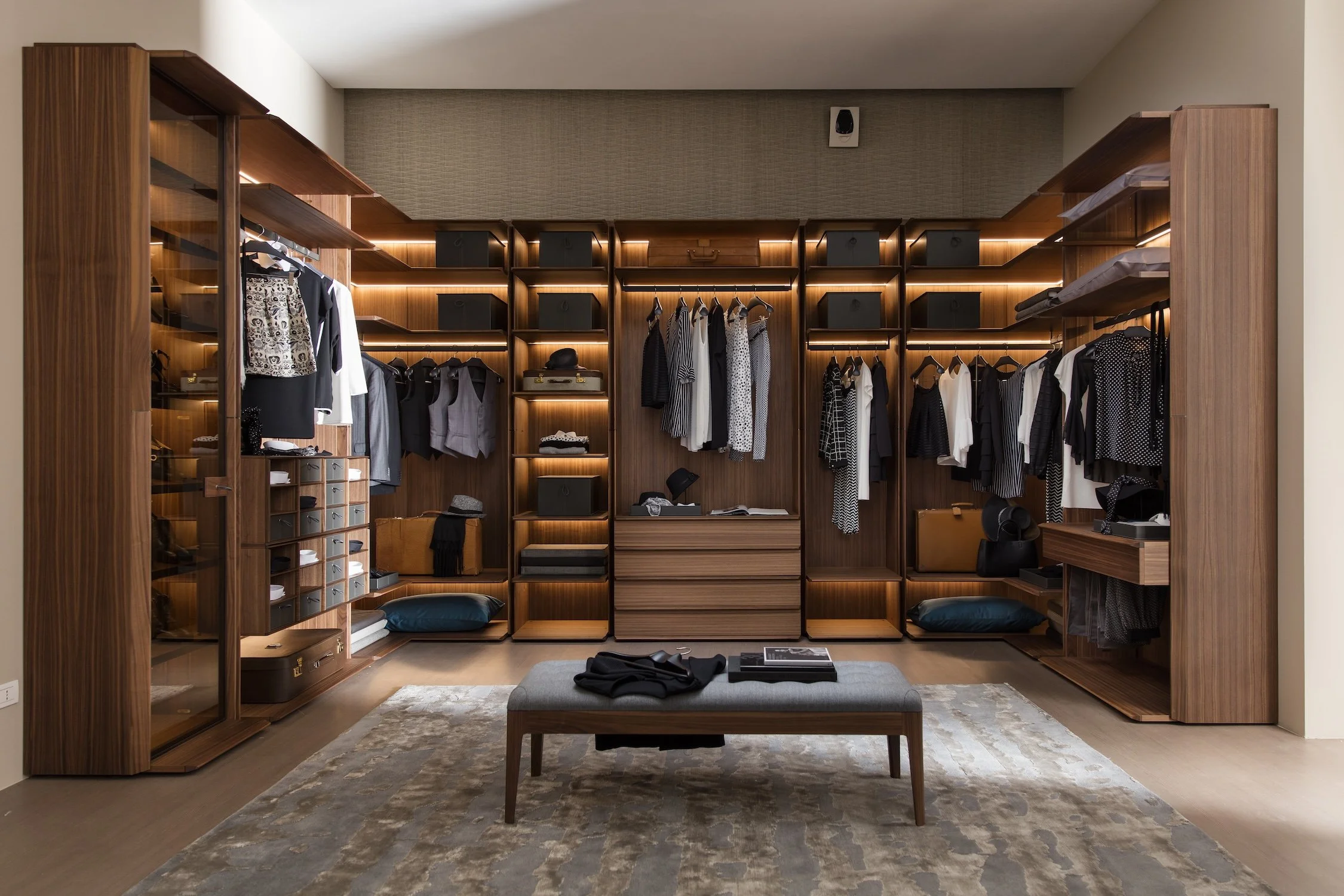 Modern walk-in closet with wooden shelves and hanging space for clothes, a bench in the center, and a patterned rug on the floor.