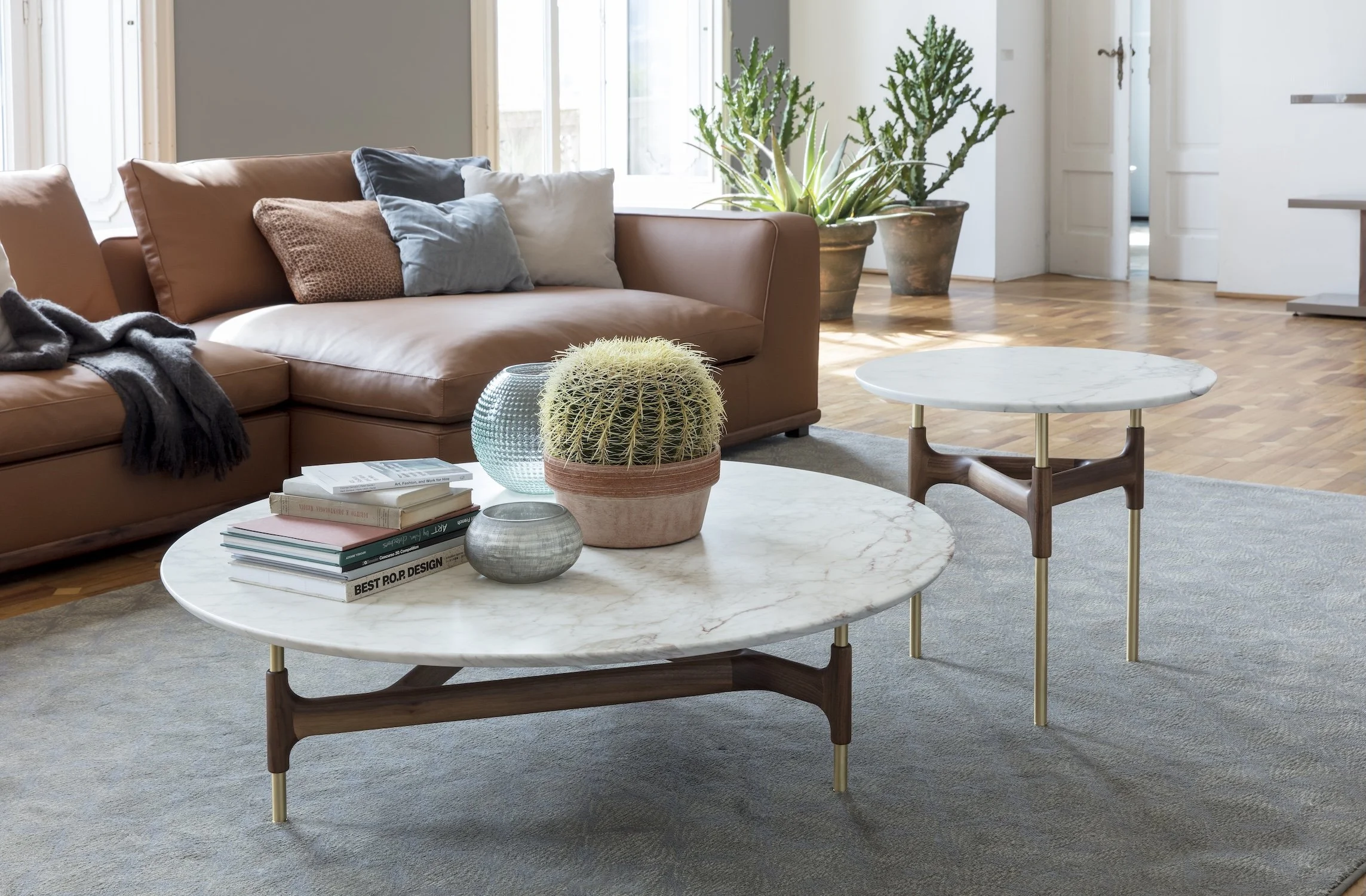 Living room with a tan sofa, throw pillows, a gray blanket, and two coffee tables with marble tops and wooden legs, decorated with books and potted plants, with a large window and hardwood floors.