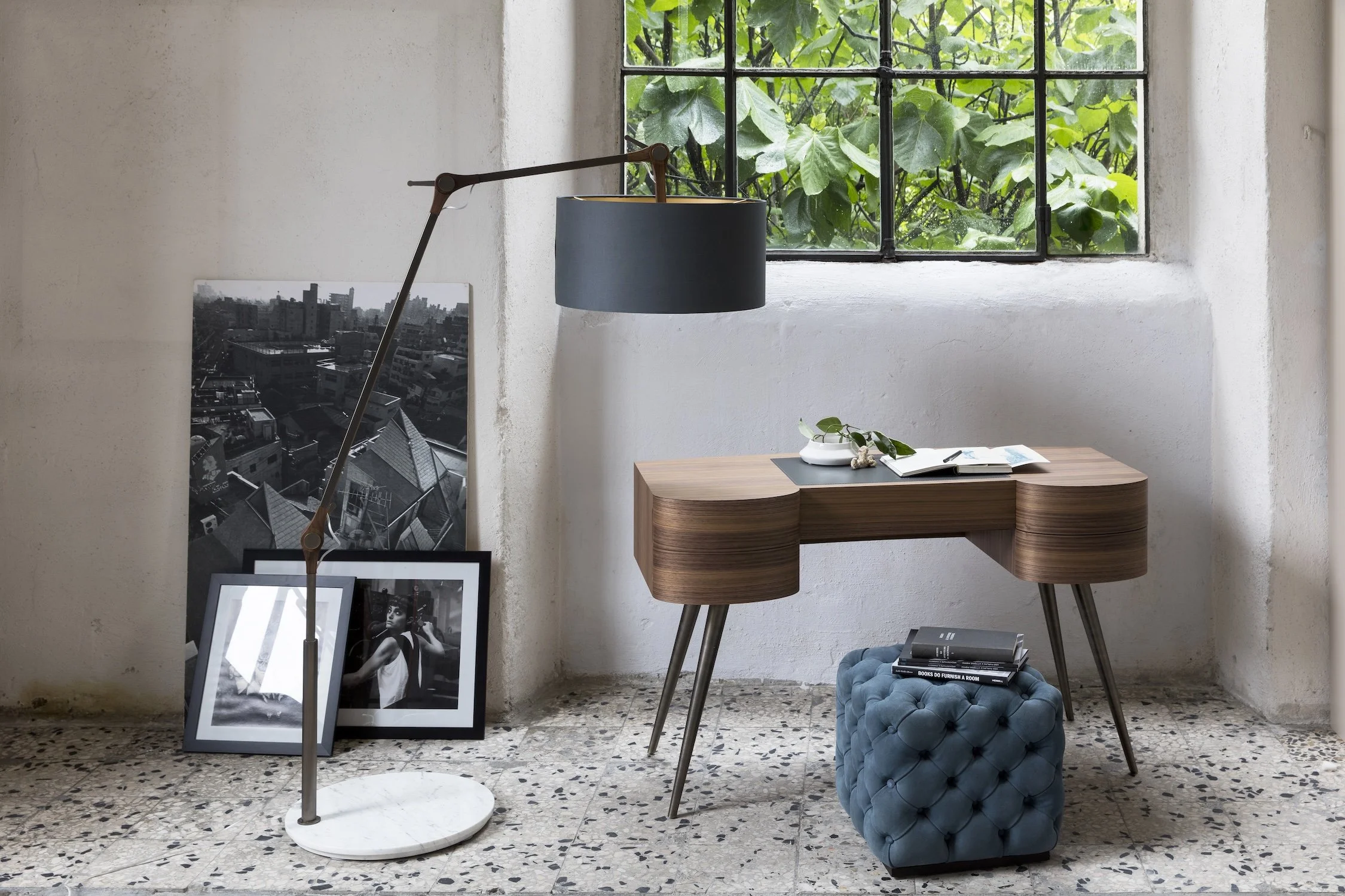 Interior space with a white wall and terrazzo floor, featuring a wooden desk with tapered legs. On the desk are an open notebook, a plant, and decorative stones. A blue tufted ottoman with books is placed underneath the desk. To the left, two framed 