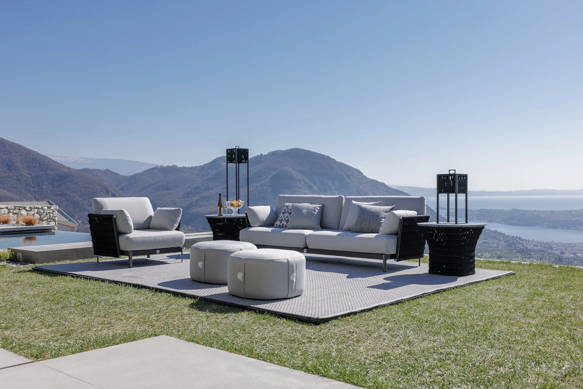 Outdoor patio seating area with white and black furniture, including a sofa, armchair, ottomans, and side tables, set on a rug with a scenic mountain and lake view in the background.