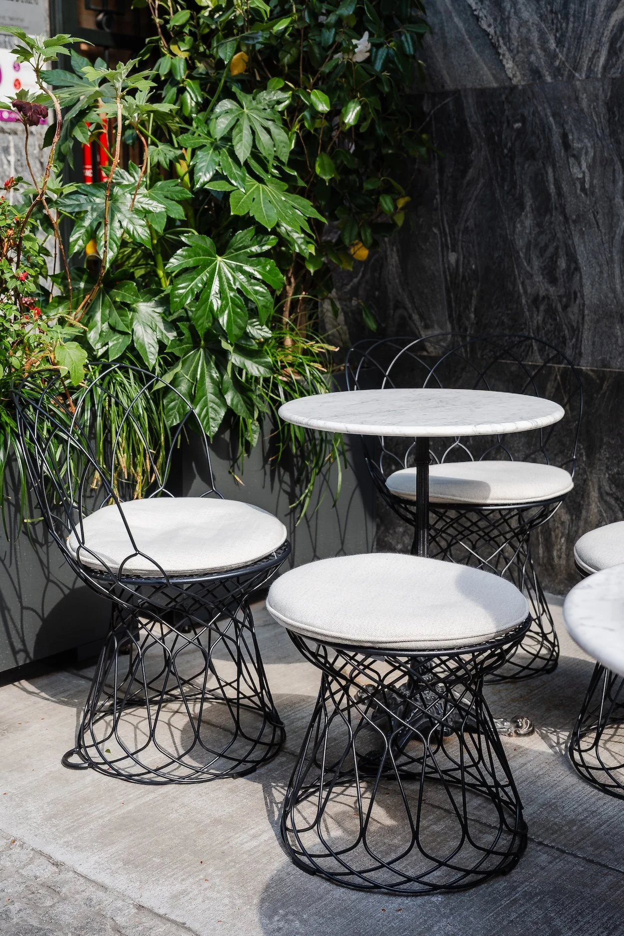 Outdoor seating area with black wire chairs with white cushions and white marble-topped tables, surrounded by green plants and a dark textured wall.