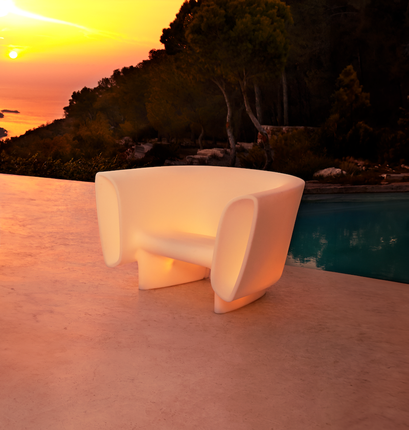 A modern, glowing white chair on a poolside patio during sunset, with trees and sky in the background.