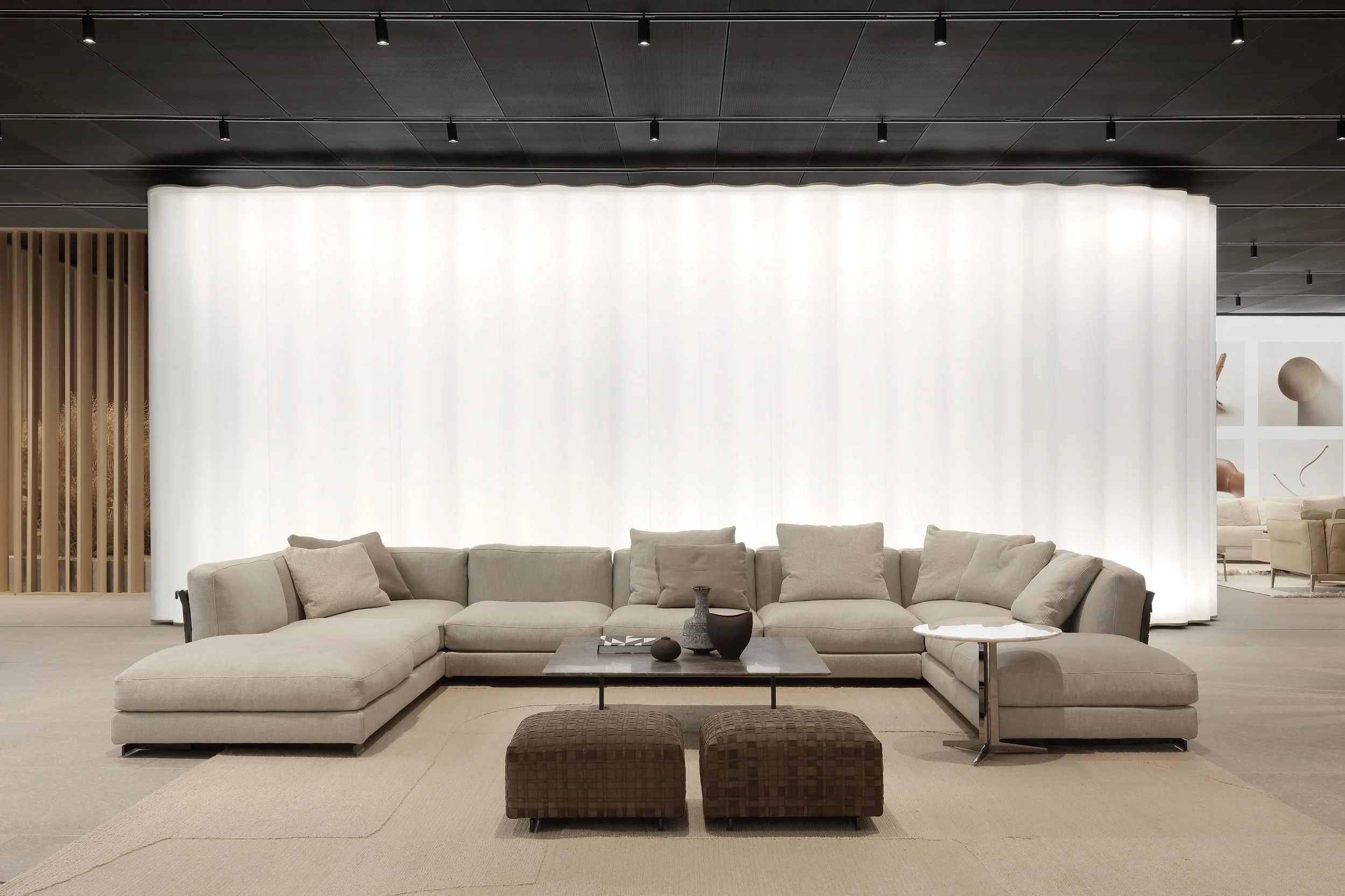 Modern living room with a large beige sectional sofa, a black coffee table with decorative vases, two brown ottomans, and a white floor-length illuminated wall panel.