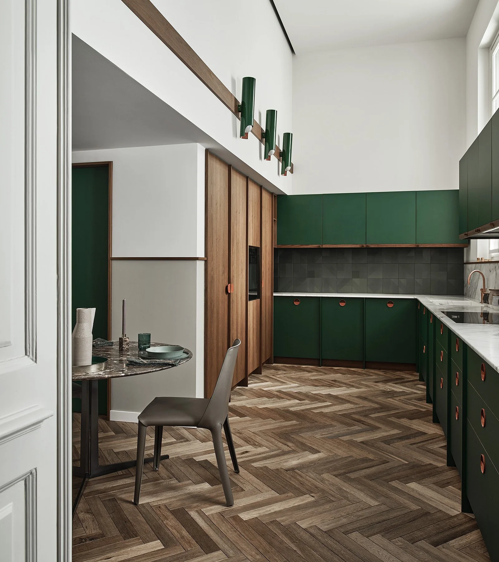 Modern kitchen with green and wooden cabinets, herringbone wood flooring, marble countertop, and black appliances. A small dining area with a round marble table, beige chair, and decorative vases.