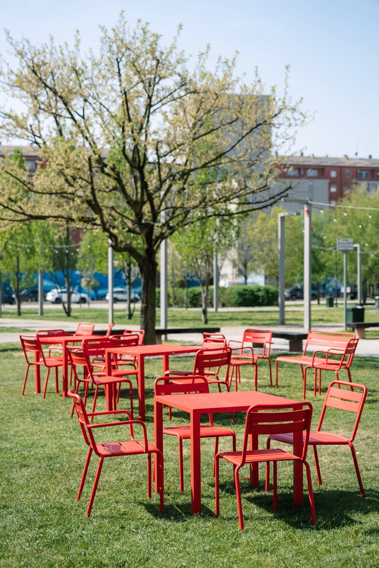 Red outdoor tables and chairs on green grass in a park with trees, benches, and buildings in the background.