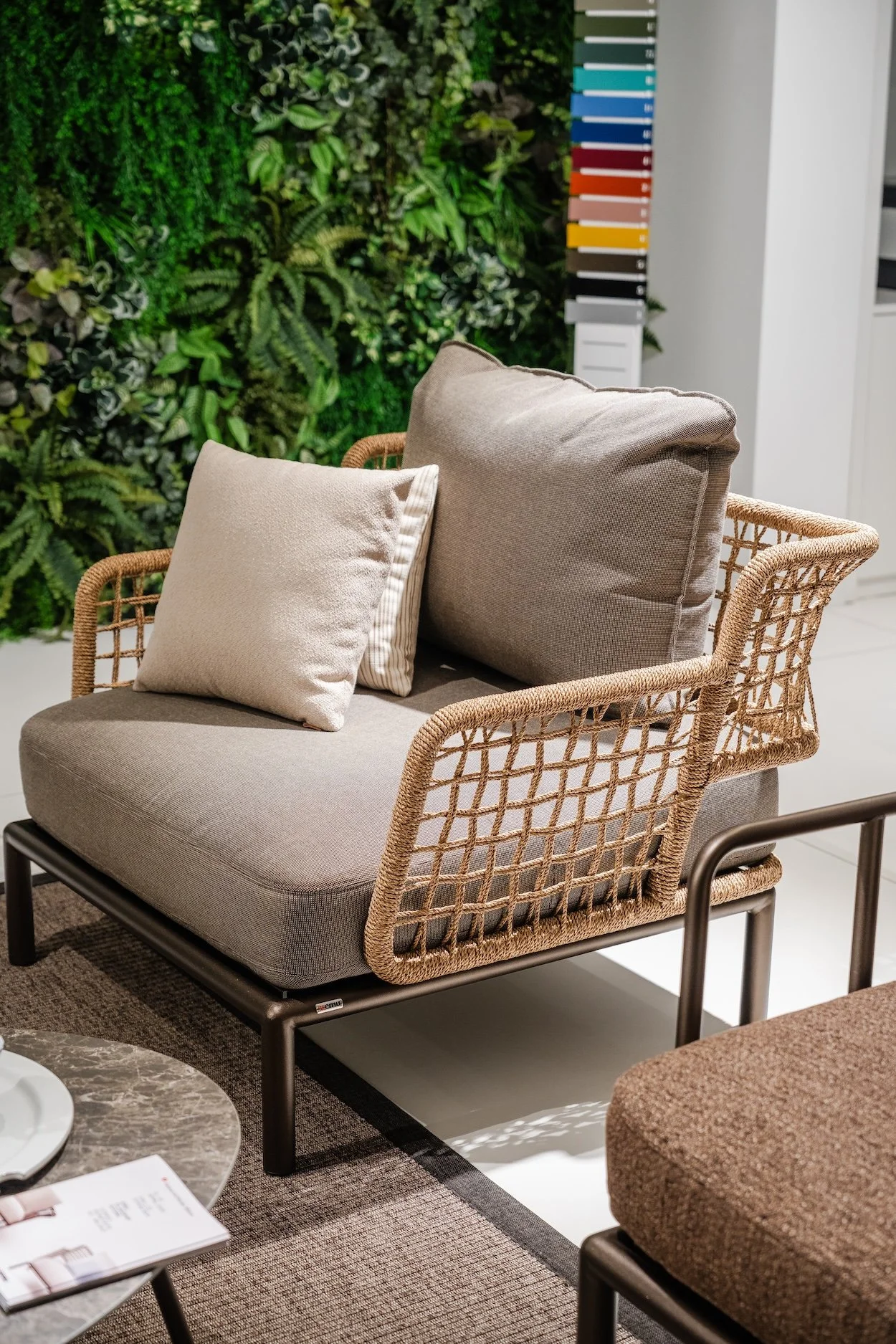 A beige cushioned loveseat with a wicker frame, two beige pillows, and a small beige striped pillow, set in a room with a green plant wall and a color palette display in the background.