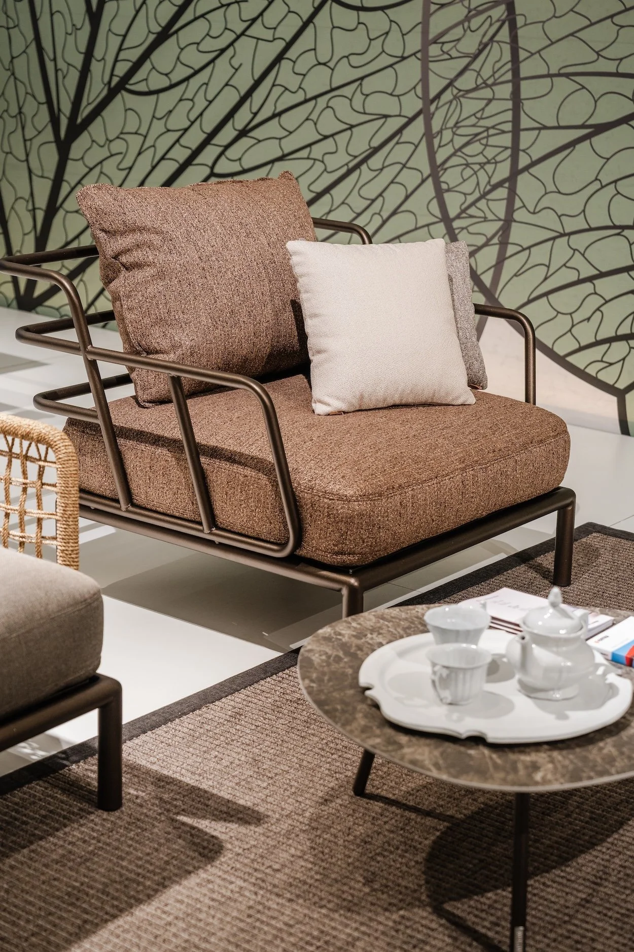 A brown sofa with two cushions, one beige and one gray, in a modern interior with a patterned green and black wall behind it. A round coffee table with a white tea set sits in front, on a brown and beige area rug.
