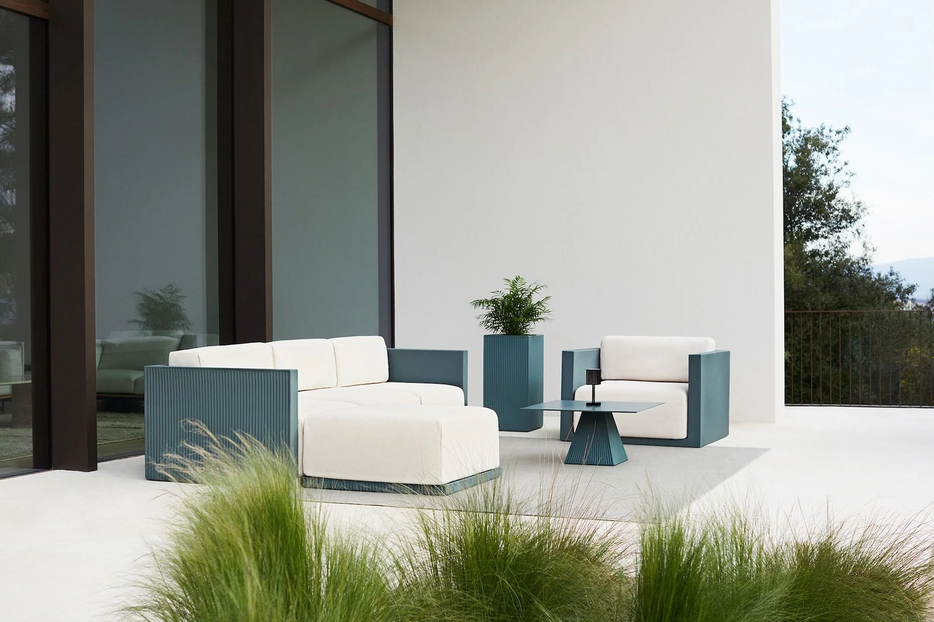 Modern outdoor patio with white and blue seating, a small black table with a candle, potted green plants, and a white wall with large glass windows.