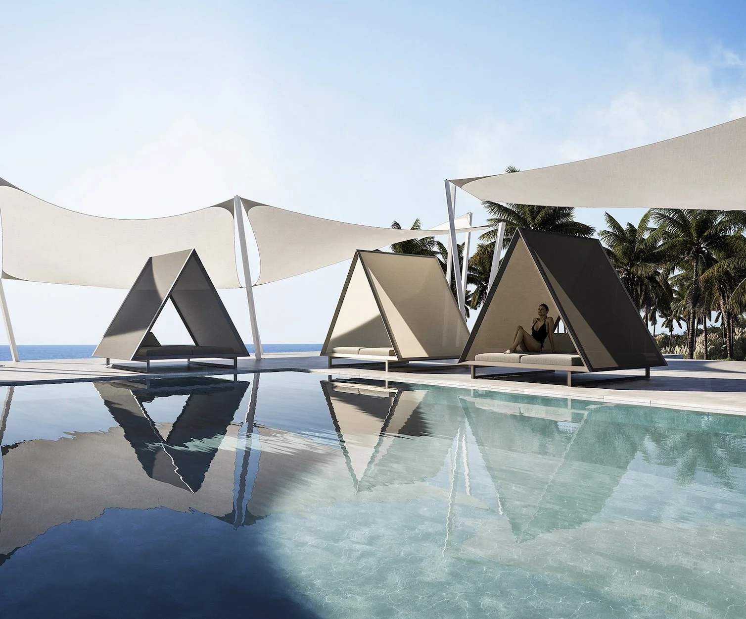 Poolside cabanas with modern triangular design and sun loungers, shaded by large white canopy sails, overlooking a beach with palm trees and the ocean in the background, and a woman sitting inside one of the cabanas.