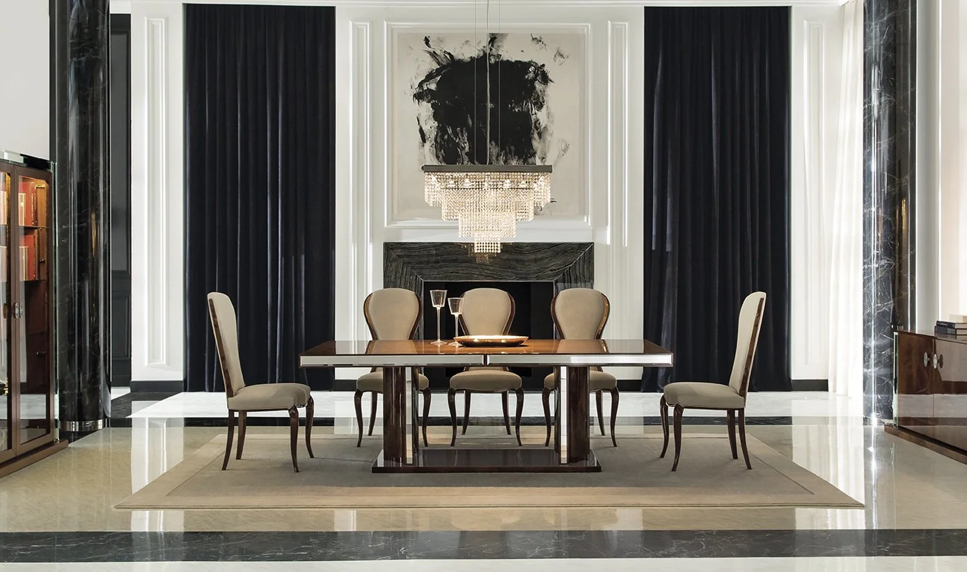 Elegant dining room with six beige upholstered chairs around a rectangular wooden dining table, a chandelier hanging above, a black abstract painting above a black fireplace, and tall dark curtains on either side of the fireplace.