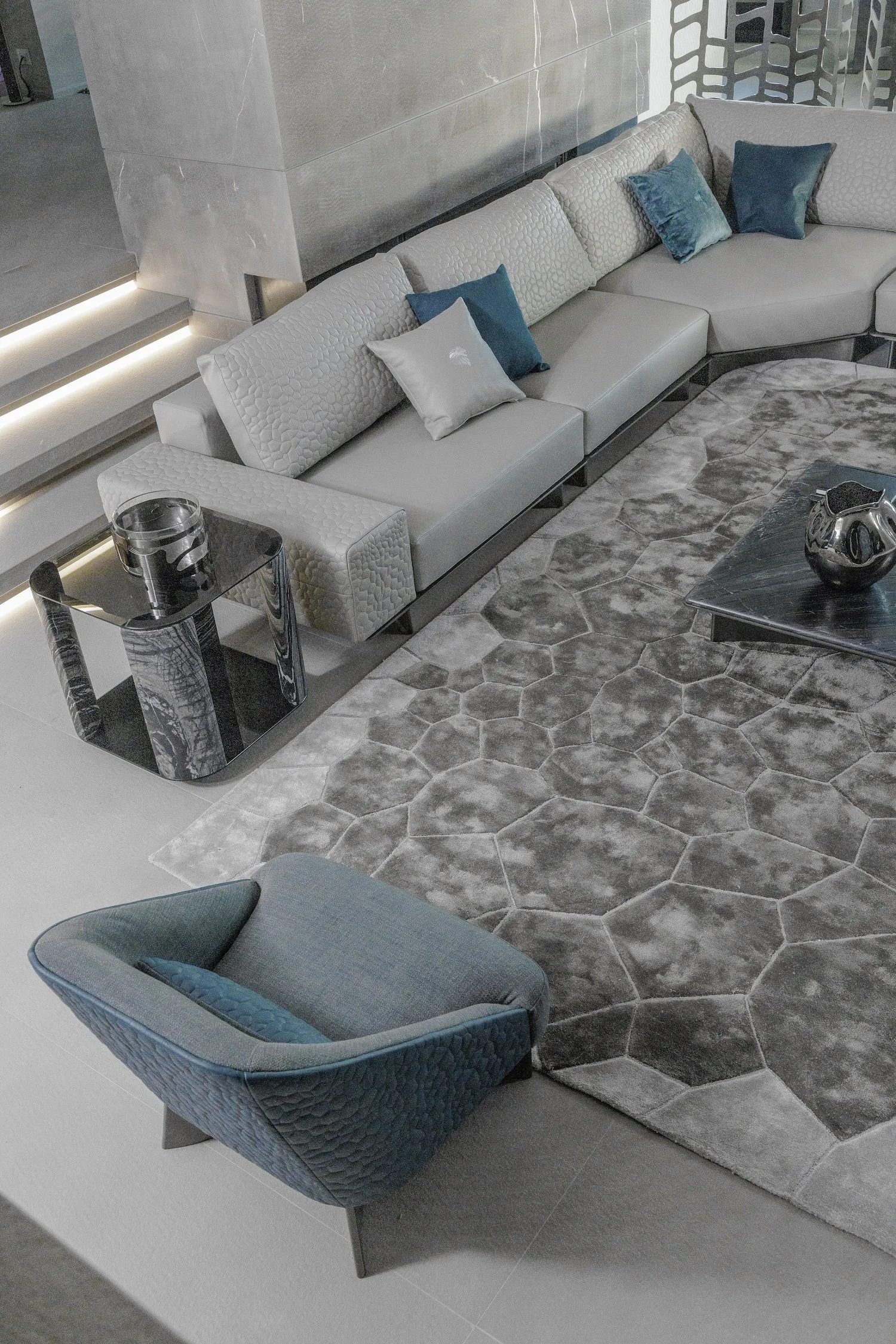 Modern living room with gray sectional sofa, a gray and blue pillows, a gray patterned armchair, a black marble side table with a glass candle holder, a gray and black hexagonal area rug, and a black marble coffee table with a metallic decorative scu