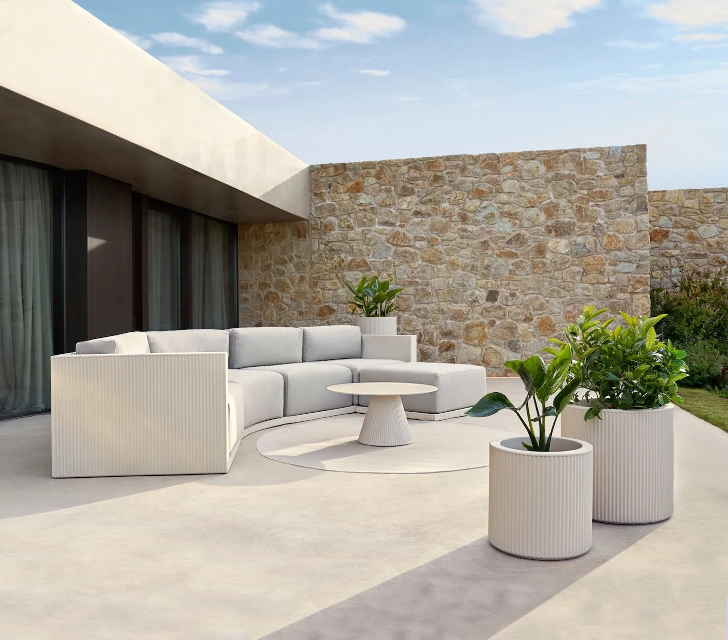 Modern outdoor patio with a white sectional sofa, round white coffee table, and three large white planters with green plants, against a stone wall and a building with large glass doors.