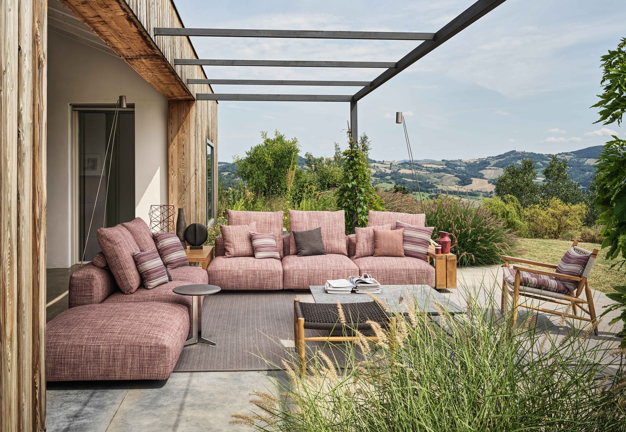 Outdoor patio with pink sofa, chairs, and side tables, overlooking a hilly landscape with trees and grass, under a clear sky.