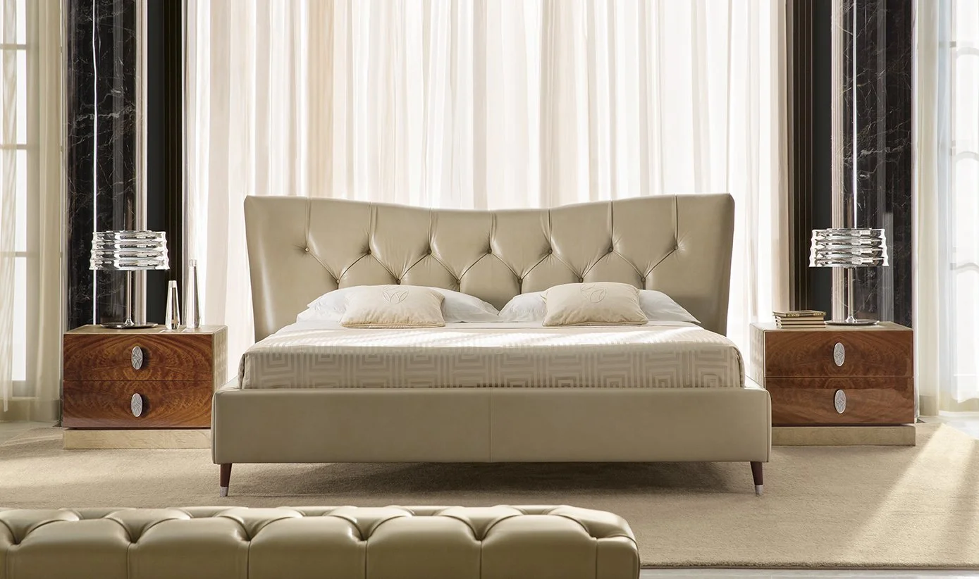 A luxurious bedroom with a beige upholstered bed, two matching bedside tables with glass lamps, and sheer ivory curtains.