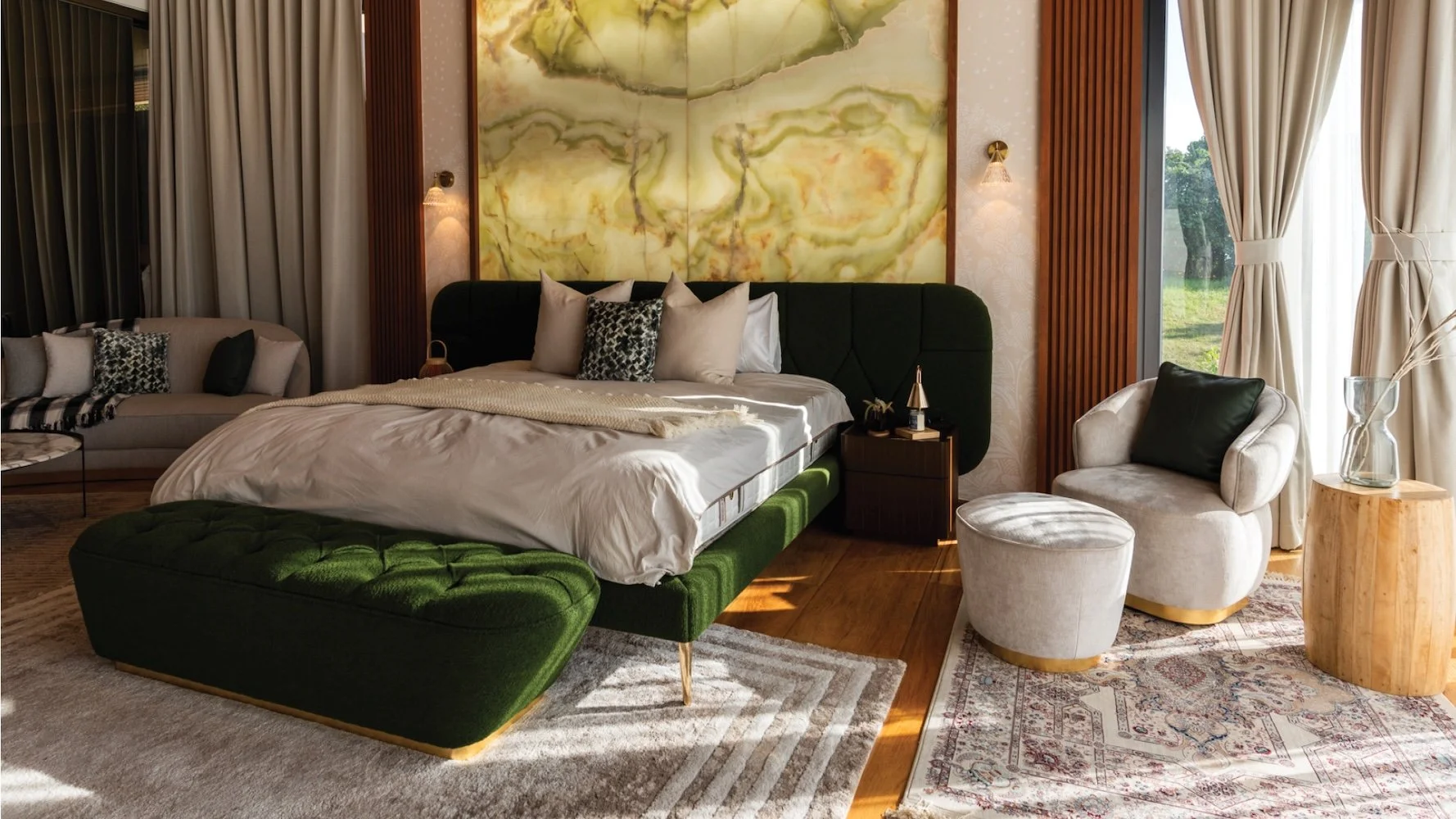 A bedroom with a large bed featuring a dark green upholstered headboard and base, decorated with multiple pillows, set against a wall with a large abstract yellow-green onyx painting. To the right, there's a white armchair with a dark green pillow, a