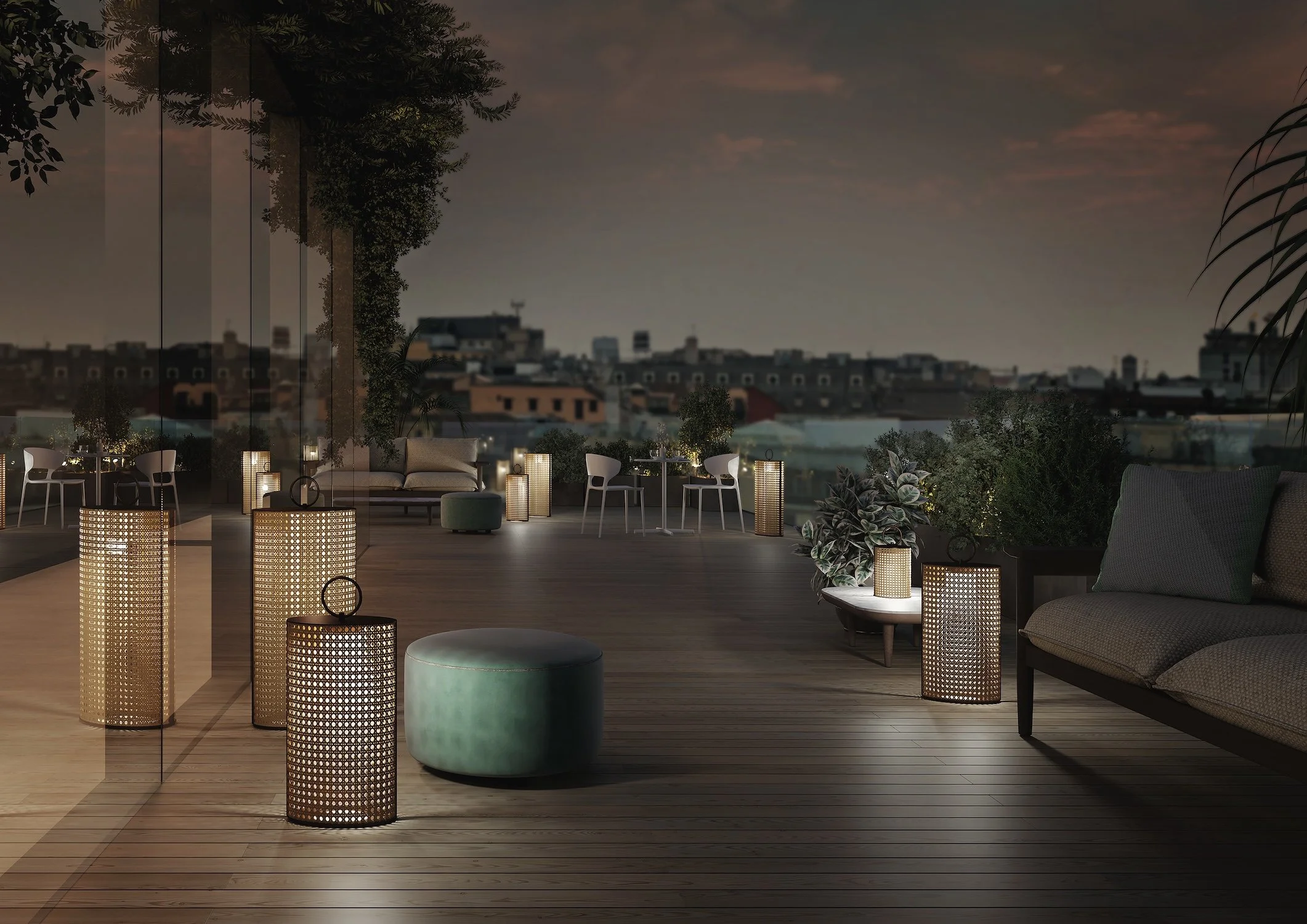 A rooftop terrace during sunset with modern outdoor furniture and illuminated lanterns, overlooking a city skyline.