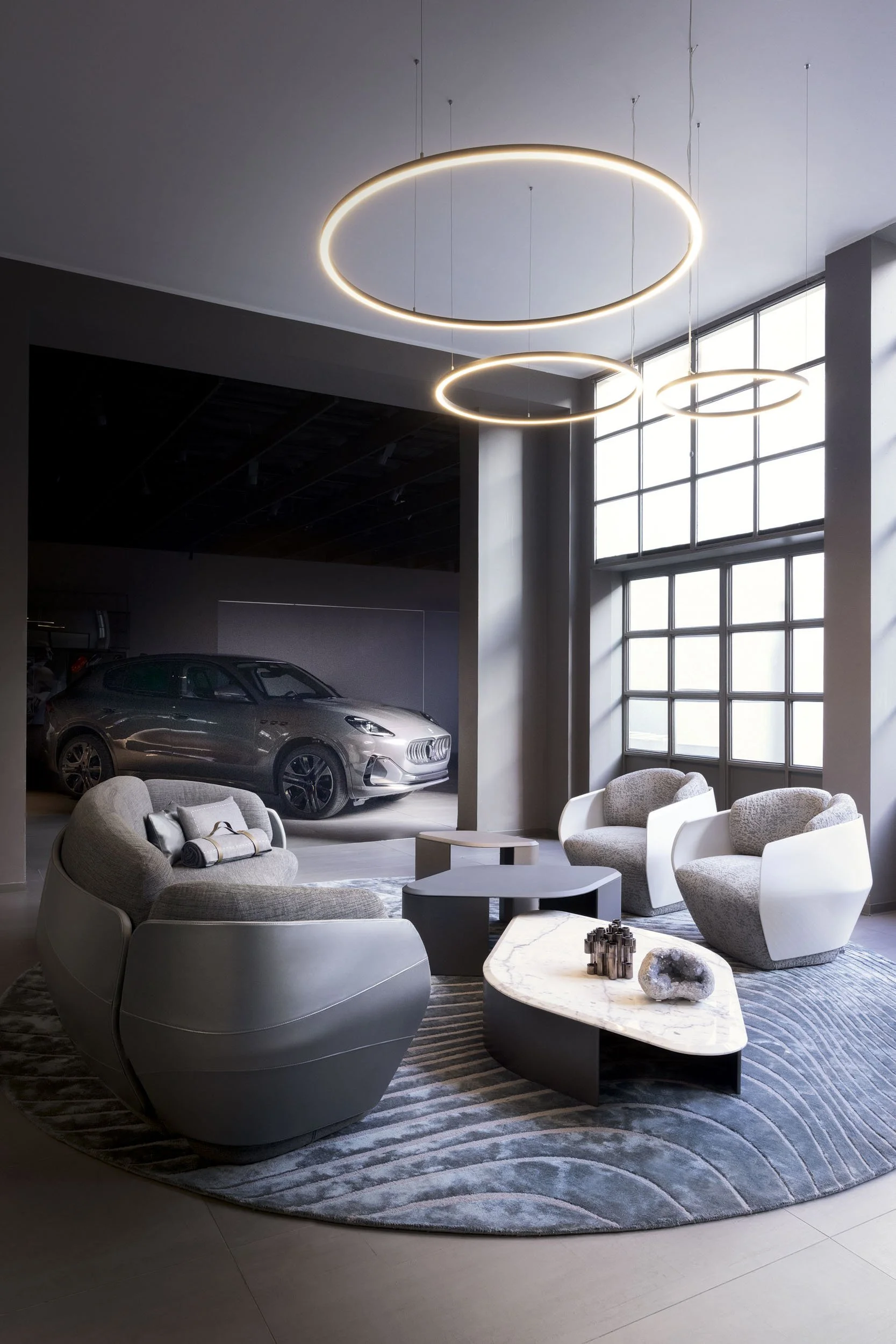 Modern interior with gray and white chairs, marble-topped coffee tables, a gray area rug, and large industrial-style windows. An electric car is parked in the background.