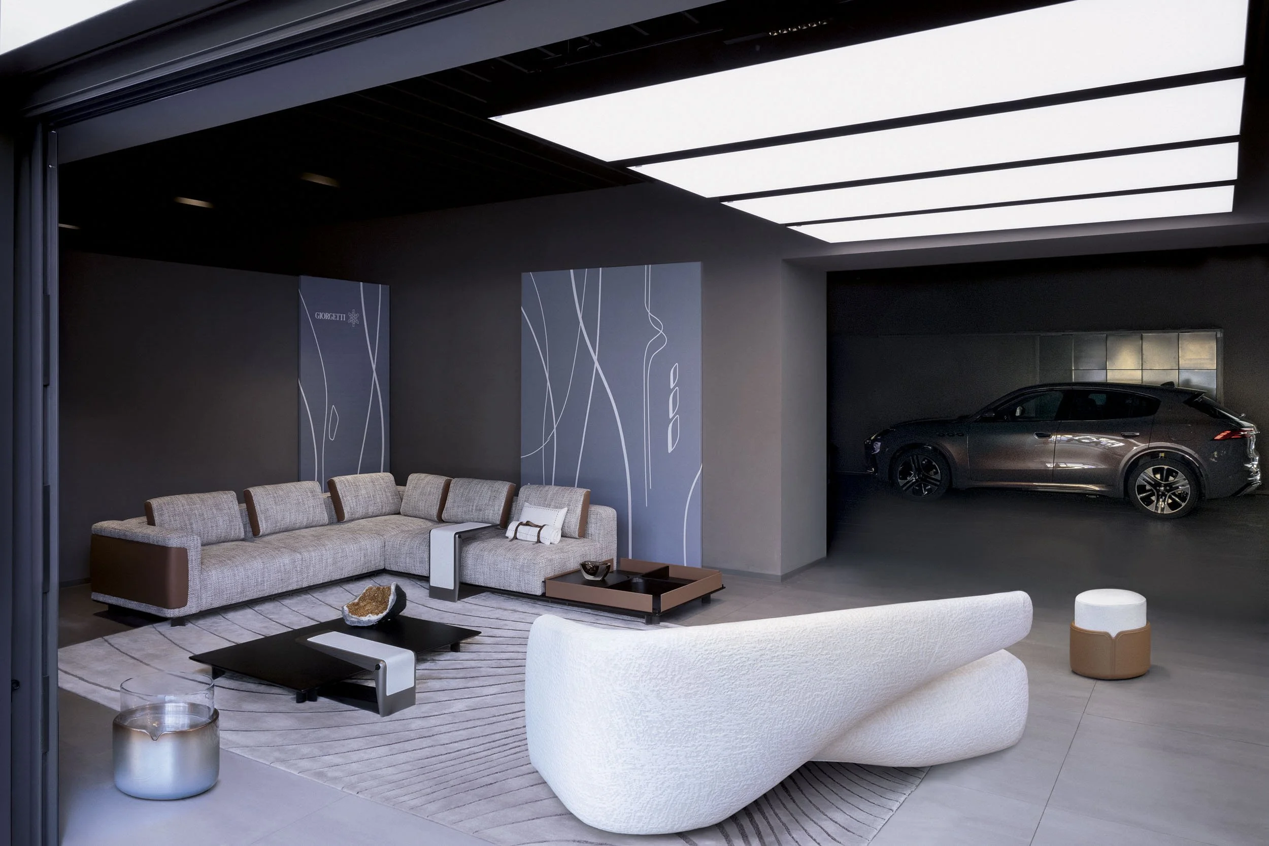 Modern living room with a large sectional sofa, a white curved lounge chair, a black coffee table, and a small white and brown ottoman, with a black car parked in a garage space adjacent to the room.