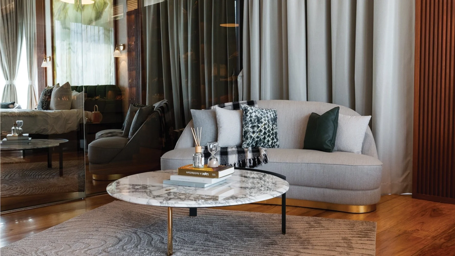Living room with a beige curved sofa, decorative pillows, and a marble coffee table with books and diffuser, separated by a glass partition from a bedroom with a bed and curtains.