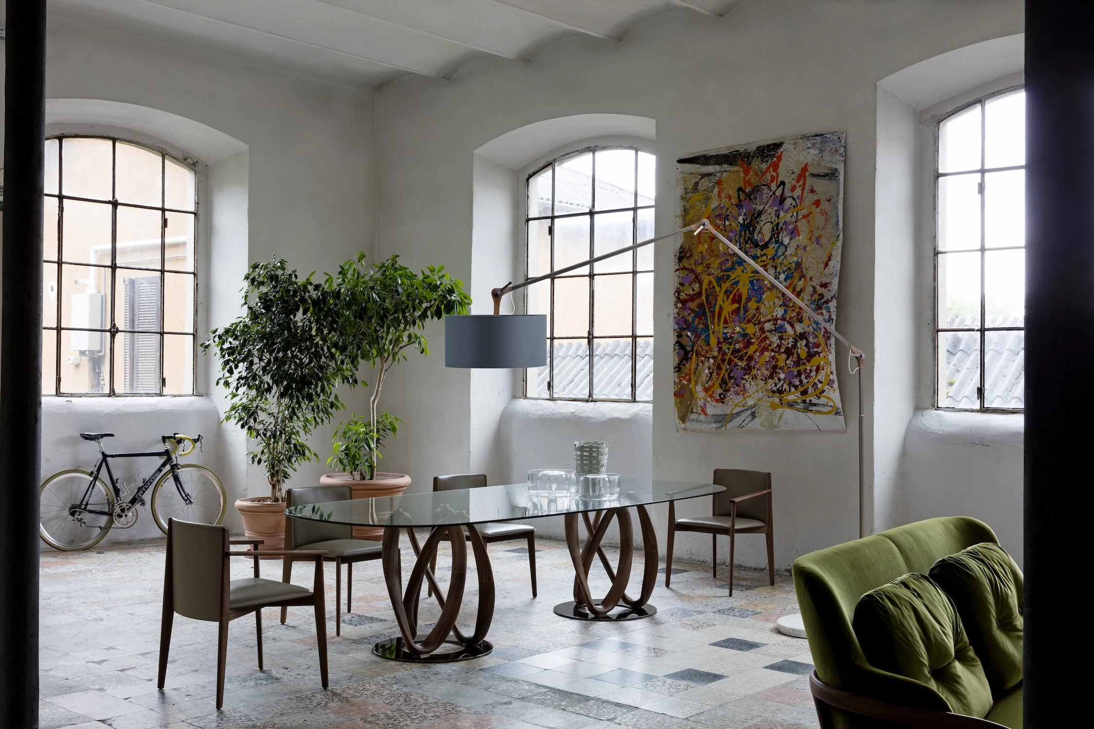Interior of a spacious room with large arched windows, an abstract painting on the wall, a glass-top dining table with decorative items, four chairs, a green sofa, a bike leaning against the wall, and a potted plant.