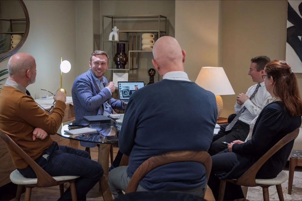 Business meeting with six people, five are seated around a table, one is working on a laptop, in a modern office setting.