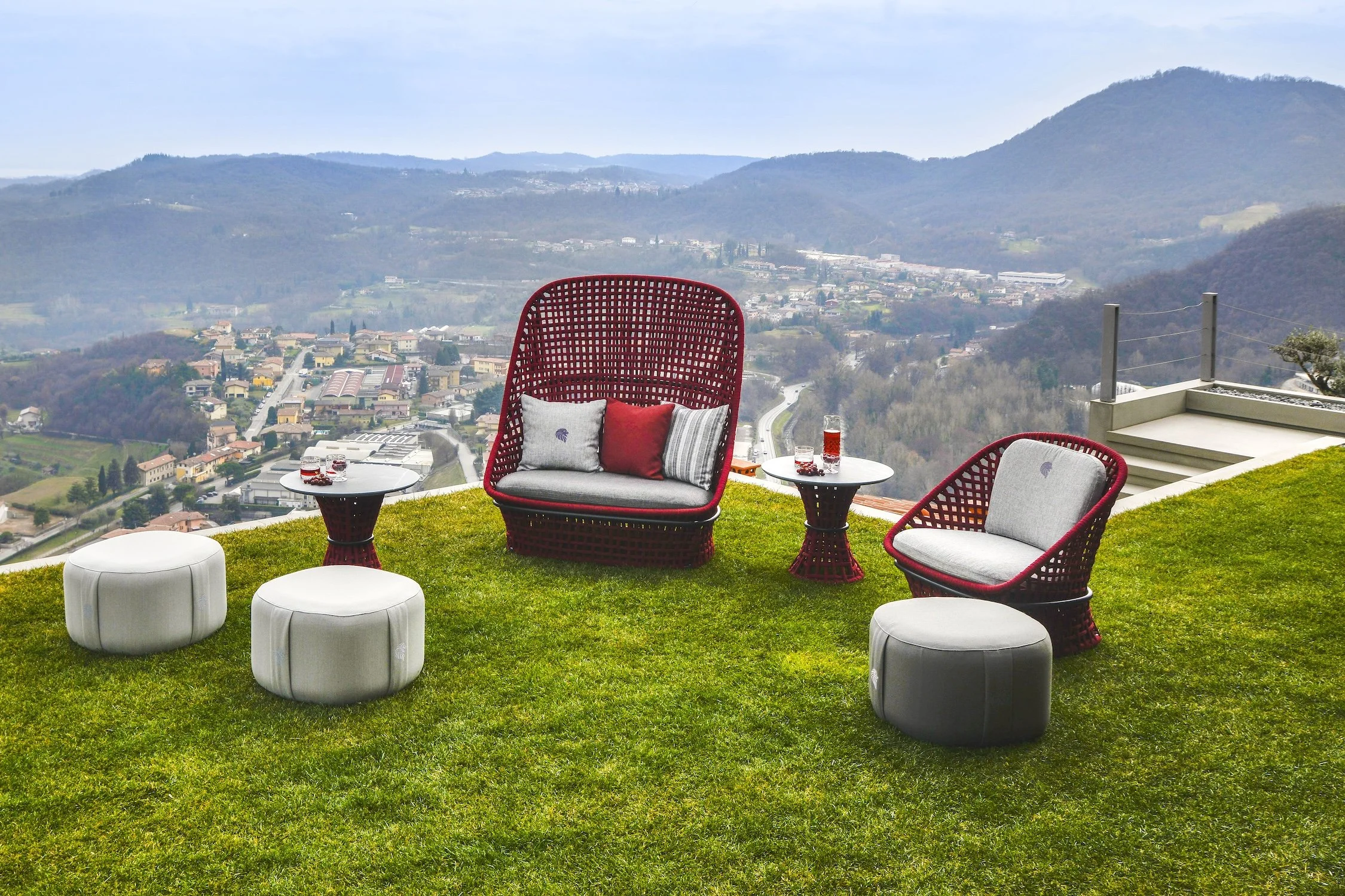 Outdoor patio furniture on a grassy hill with a view of a valley and mountains in the background. The furniture includes a large red wicker chair with pillows, two white ottomans, and two small round side tables with drinks.