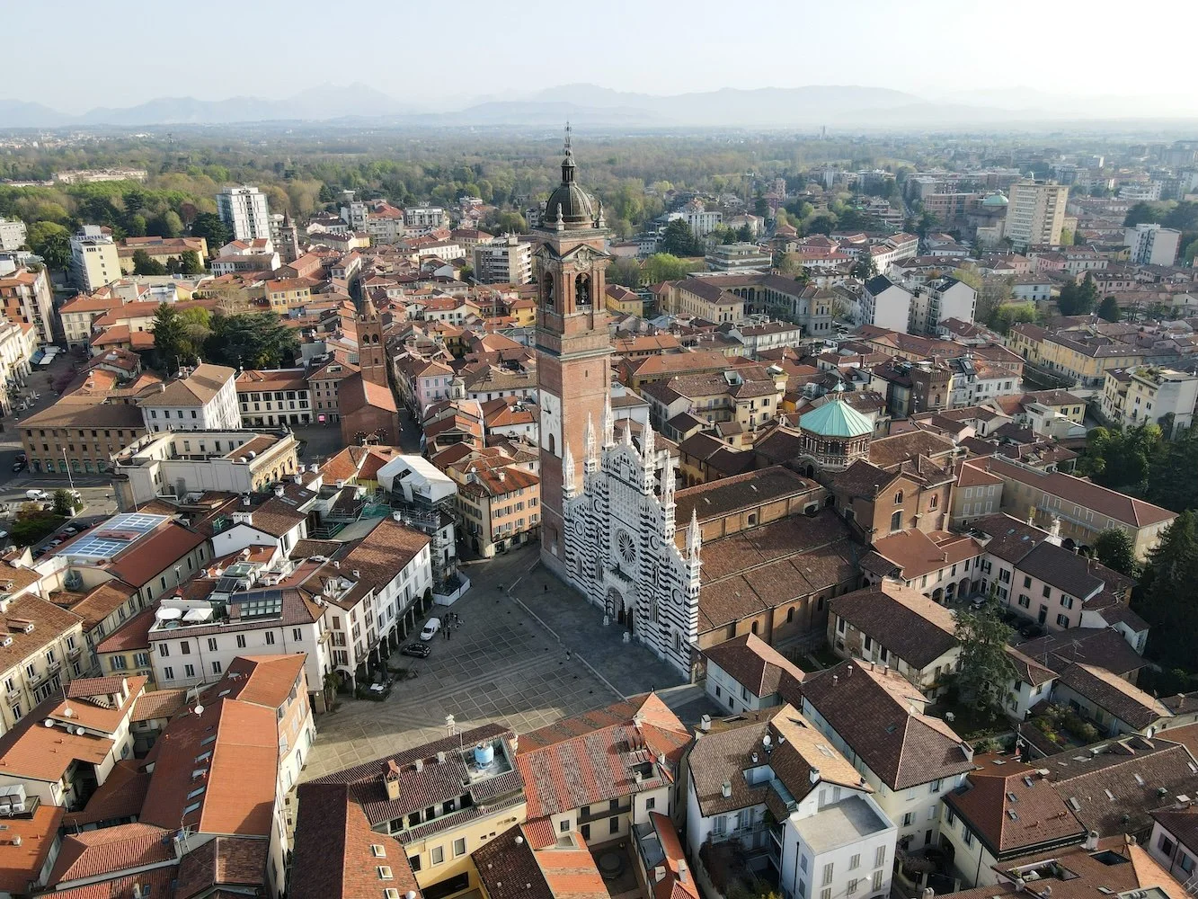 An aerial view of an Italian city with a large cathedral in the center, featuring a striped white and black facade and a tall brick clock tower. The cityscape includes numerous red-tiled roofs, small buildings, and narrow streets, with green hills and mountains visible in the distance.