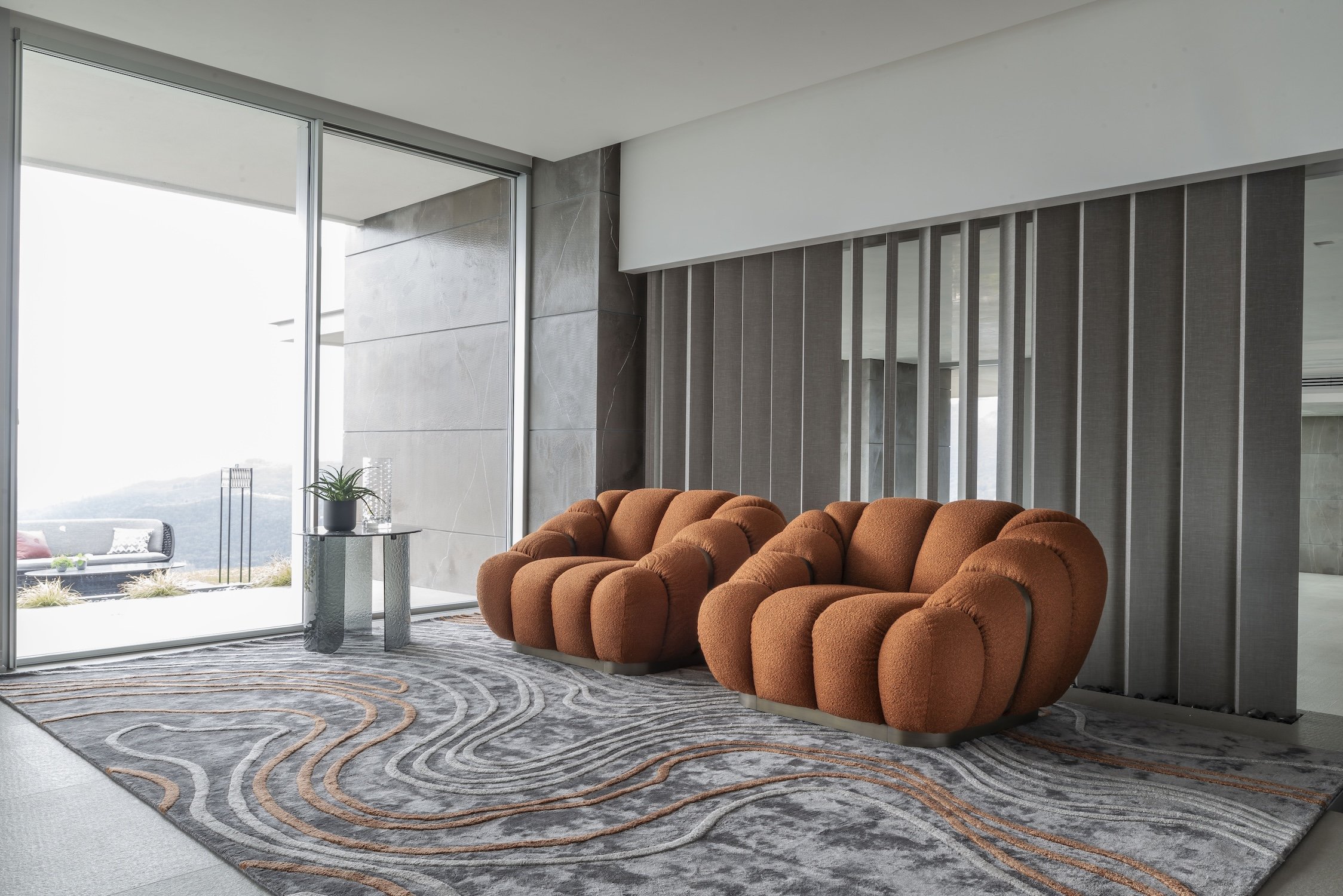 Modern living room with two orange plush armchairs, a small side table with a potted plant, and large floor-to-ceiling windows showing an outdoor patio with seating and plants.