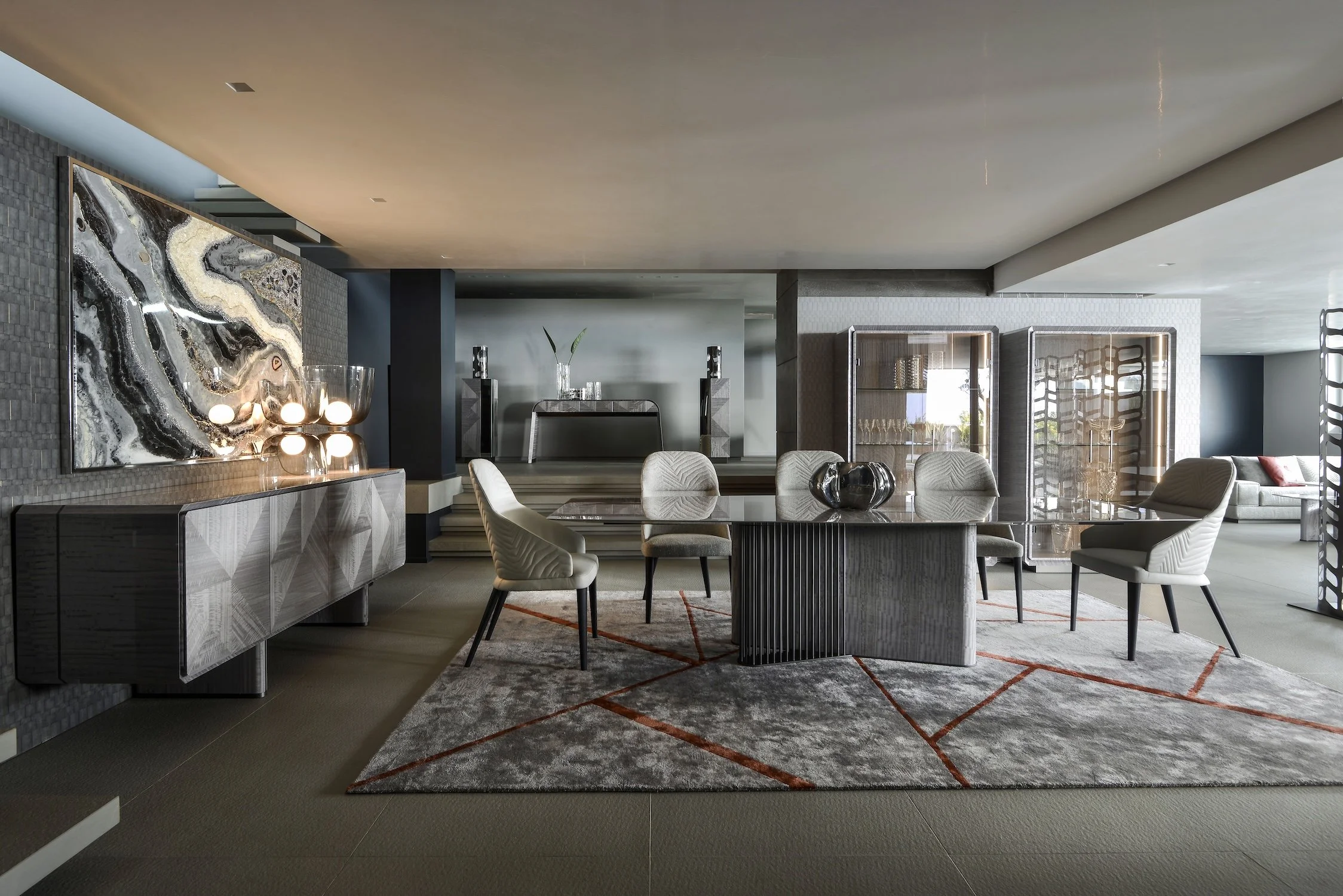 Modern living and dining area with a rectangular glass table, six upholstered chairs, a patterned area rug, a sideboard with lamp, and a TV console with a decorative sculpture on top.