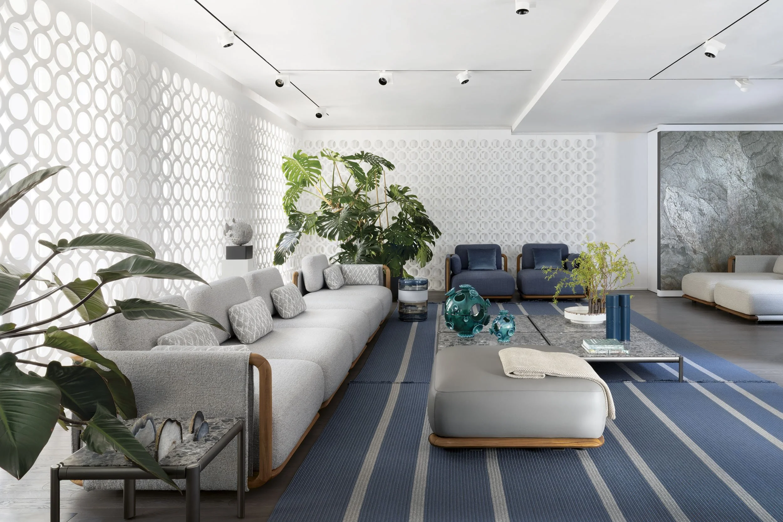 Modern living room with white walls, large gray sofa with patterned cushions, blue armchairs, large green plant, decorative blue glass objects, and a gray textured ottoman with a throw blanket, on a blue striped rug.