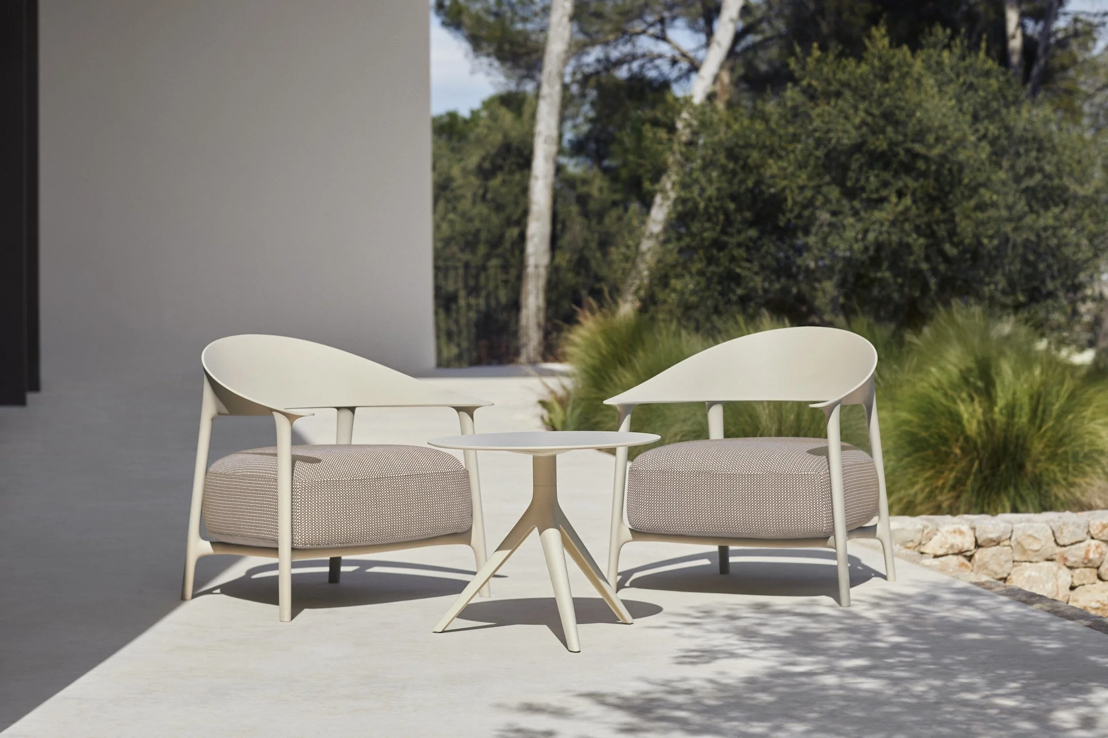 Outdoor patio with two modern white chairs with beige cushions and a small oval white table, surrounded by green bushes and trees.