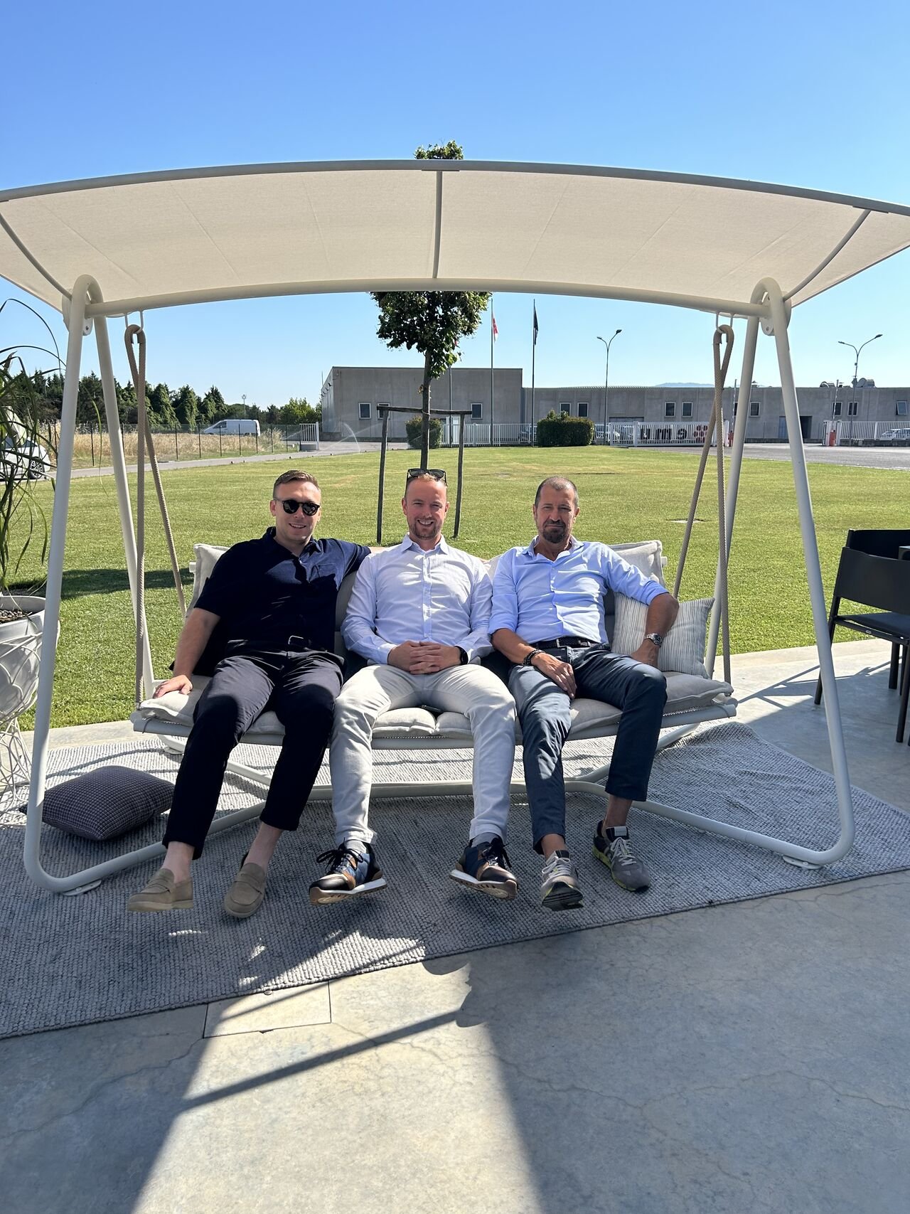Three men sitting on a large outdoor swing with a canopy, on a patio with grass and a building in the background.
