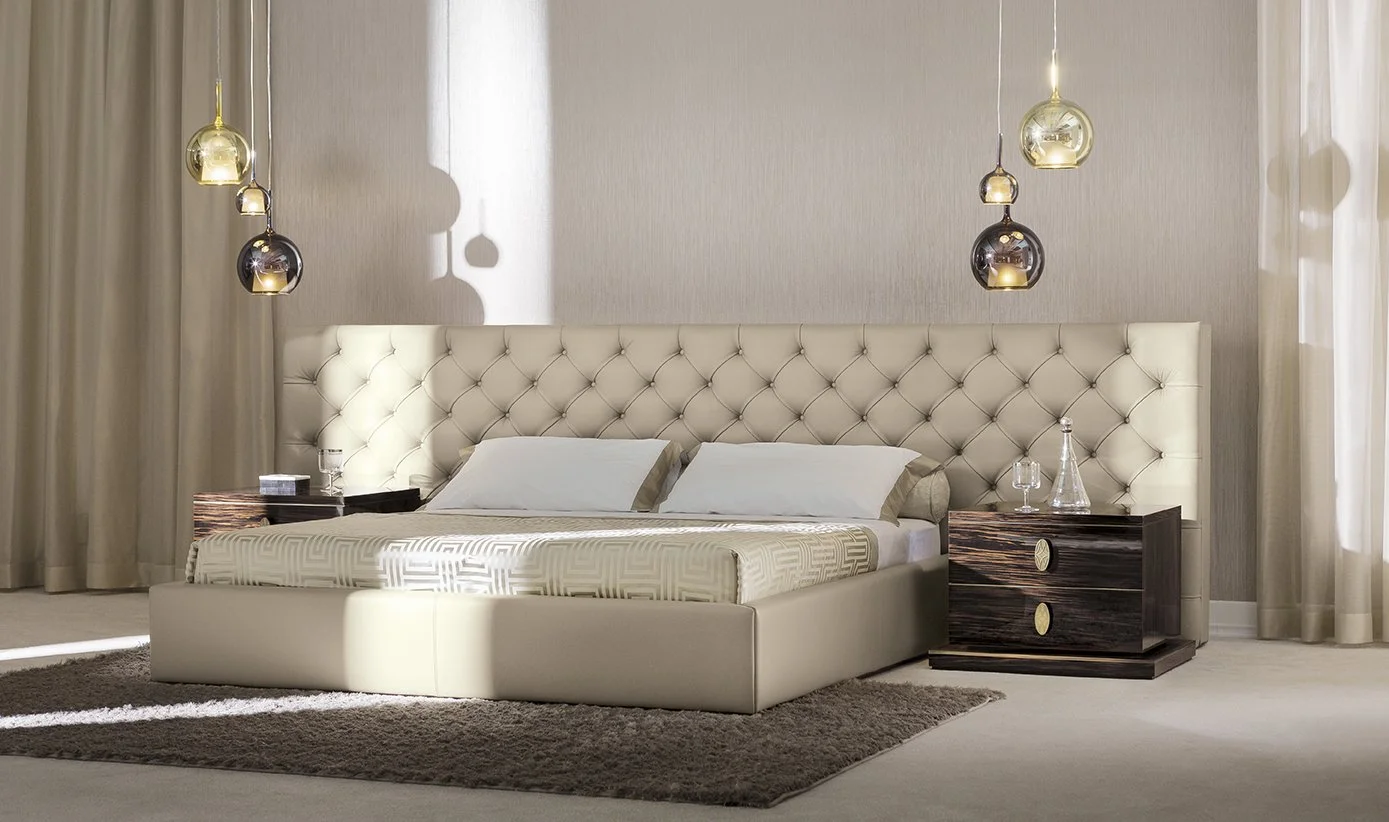 A modern bedroom featuring a large cream-colored, tufted headboard, a bed with white pillows and a patterned bedspread, and two dark wooden nightstands, each with glassware. Hanging pendant lights add a stylish touch to the space.