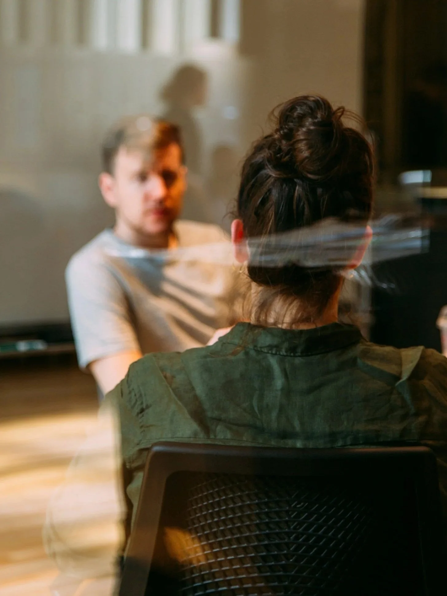 A woman with dark curly hair tied up, seen from behind, sitting at a table facing a man with short light brown hair, wearing a light-colored shirt, indoors with a blurred background.